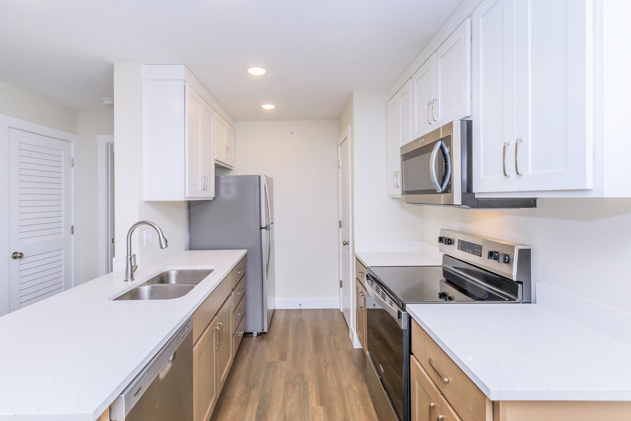 Modern kitchen featuring white shaker cabinets, stainless steel appliances including a refrigerator, oven, and microwave, and a spacious countertop with a double sink. The flooring is a warm wood tone, and there are bright overhead lights illuminating the area. The layout is open and inviting.