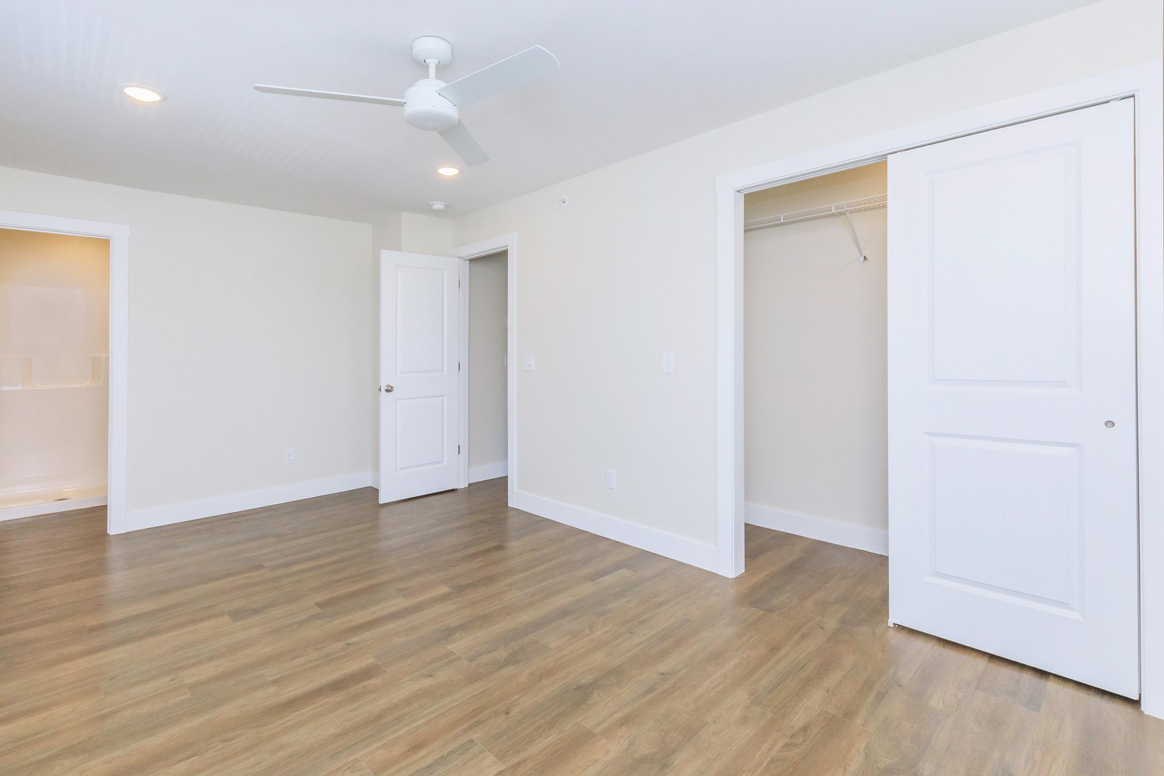 Empty room featuring light-colored walls, a ceiling fan, and wooden flooring. There are two doors: one leading to a closet and another likely leading to a bathroom or hallway. Natural light is coming from an unseen window, illuminating the space.