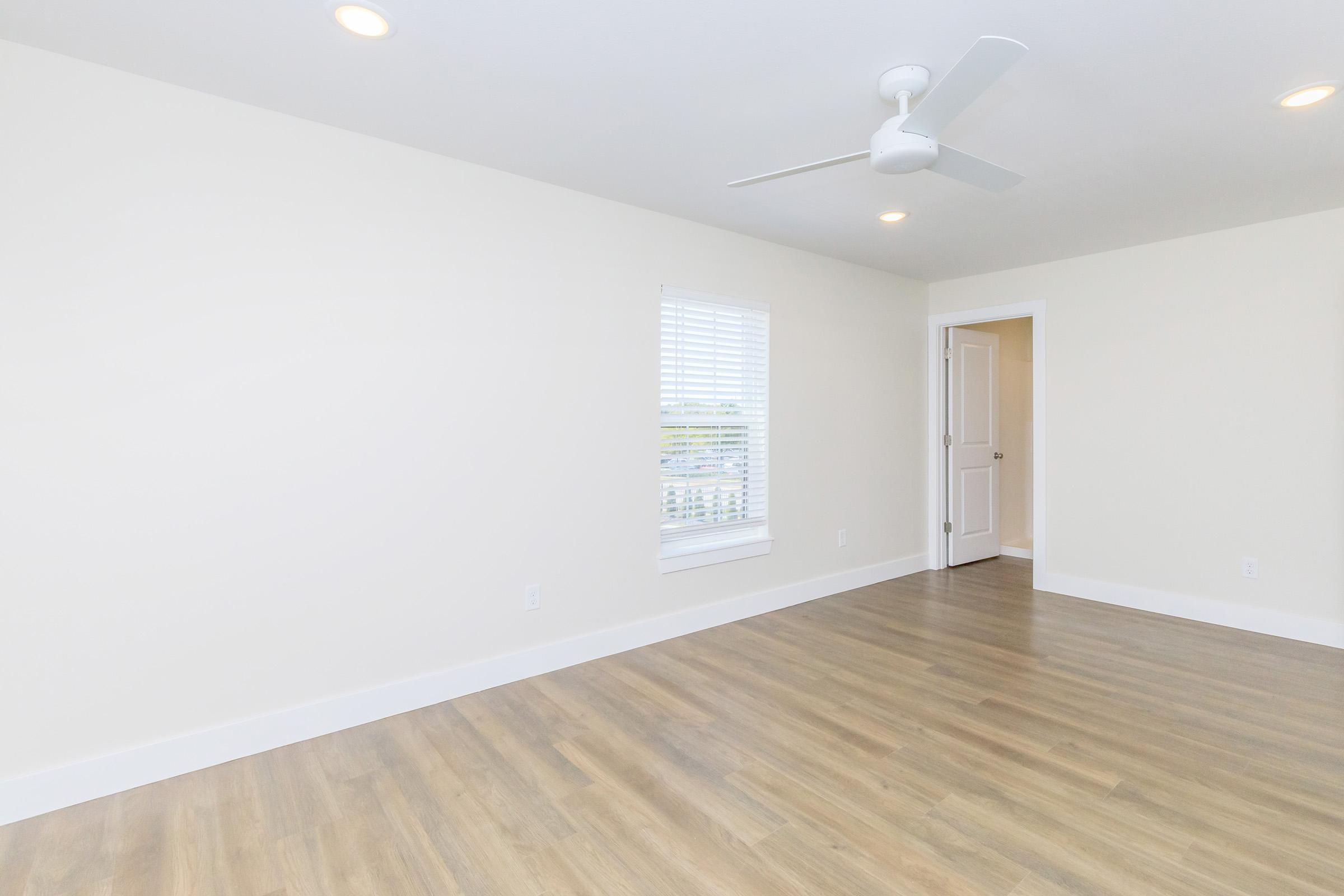 Spacious, empty room with light-colored walls and a modern ceiling fan. There’s a window with blinds allowing natural light and a door leading to another area. The floor is wooden, giving the room a warm feel, ideal for various interior design possibilities.