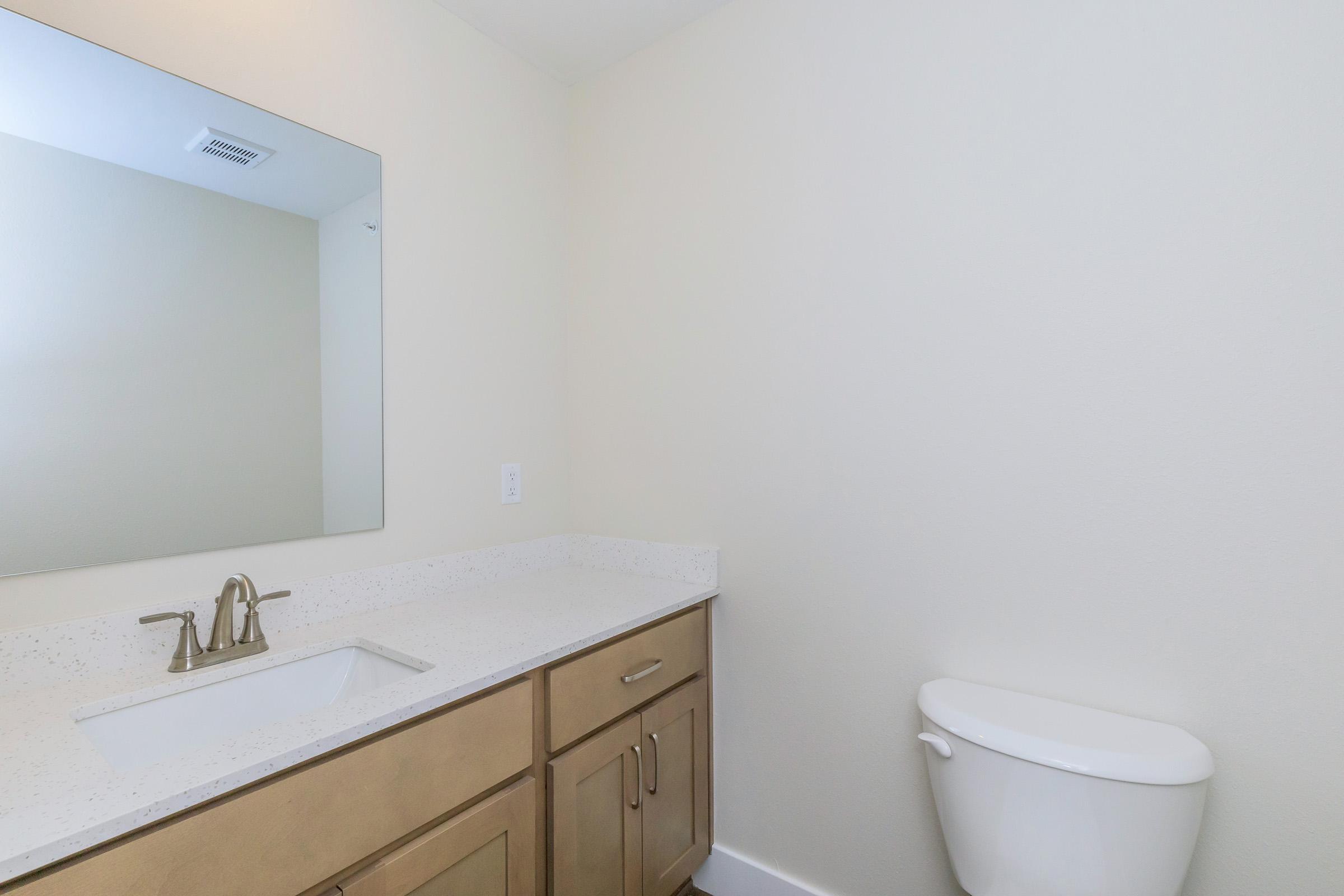 A clean, modern bathroom featuring a large mirror above a white sink with a beige countertop. On the right, there is a white toilet against a light-colored wall, contributing to a minimalist design with neutral tones.