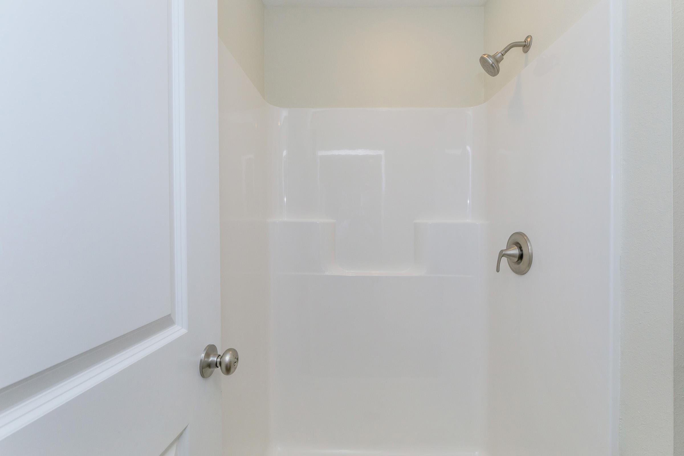 A clean, empty shower space featuring white plastic walls, a showerhead on the left, and a simple control knob. The setting is bright and minimalistic, with a focus on functionality. The door to the shower is closed, emphasizing the interior's simplicity and modern design.