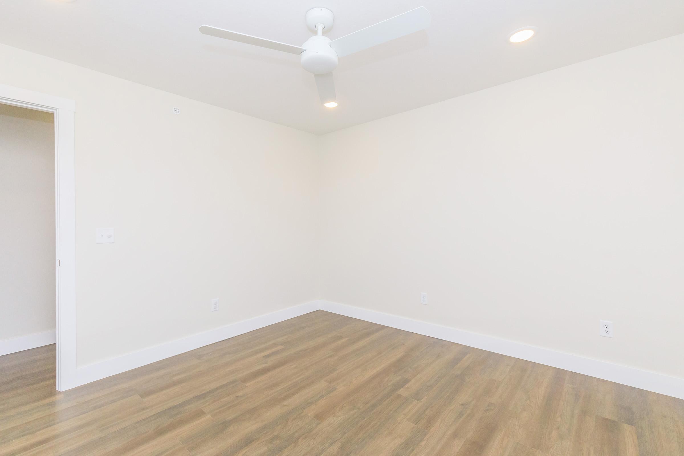 A brightly lit, empty room featuring light-colored walls, a ceiling fan, and laminate flooring. The space has a simple, modern design with no furniture, allowing for versatile use. A doorway is visible on the left side of the image.