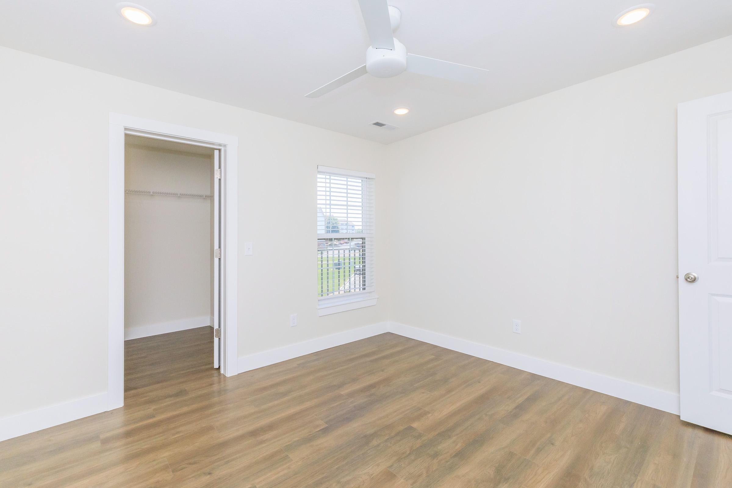 A bright, empty room with light-colored walls and wooden flooring. It features a ceiling fan and a window with white blinds. A closet is visible to the left, providing additional storage space. The overall atmosphere is clean and modern, suitable for various uses like a bedroom or office.