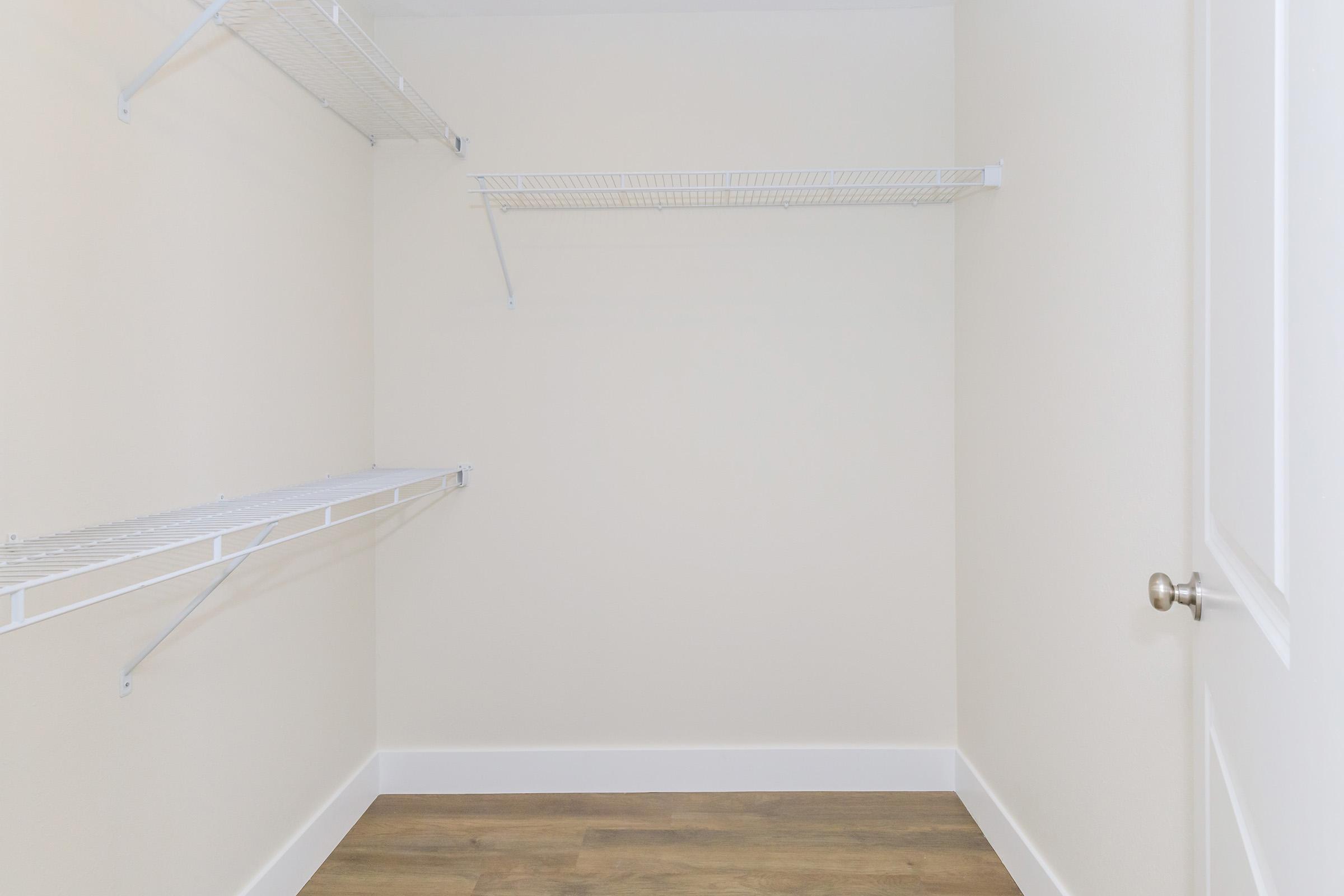 Empty closet interior with white wire shelving on two walls, light-colored walls, and a wooden floor. The closet has a door handle visible, and there's a sense of spaciousness due to the absence of items.