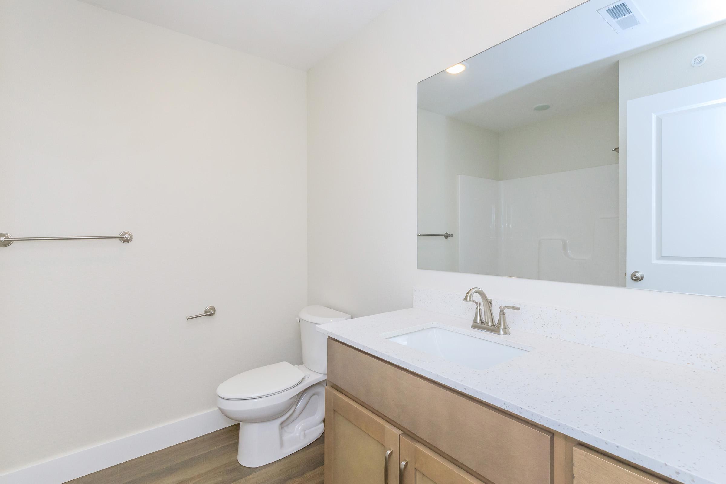 A clean and modern bathroom featuring a white vanity with a sink, a large mirror, a toilet, and a shower area. The walls are painted in a light color, and there is a towel bar next to the toilet, enhancing the minimalist design of the space.