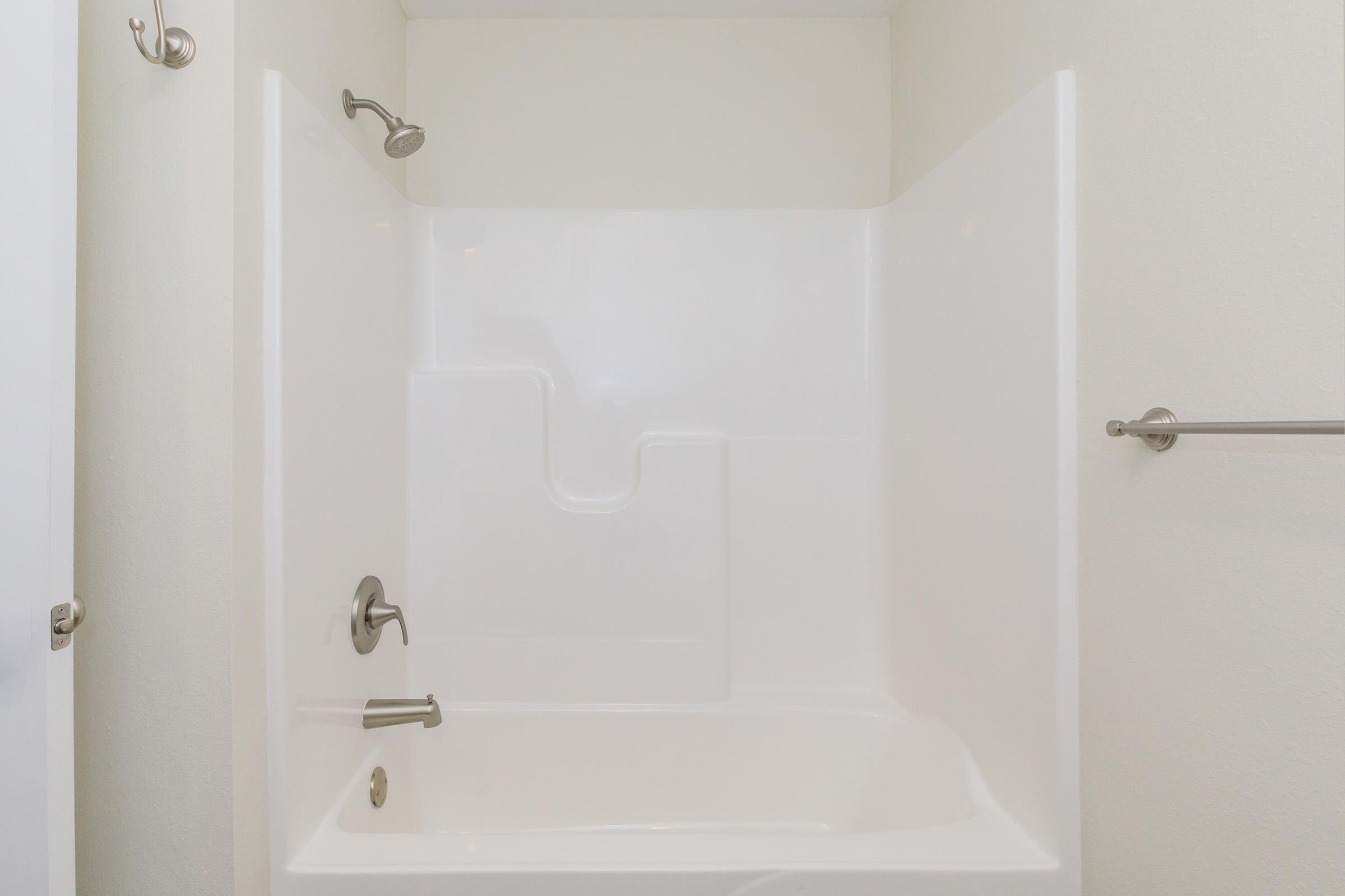 A clean, white bathtub and shower enclosure with a modern design. The tub features a built-in step and a single faucet with temperature control. The wall has a towel bar, and there is a showerhead above. The walls are painted in a light, neutral color, enhancing the brightness of the space.