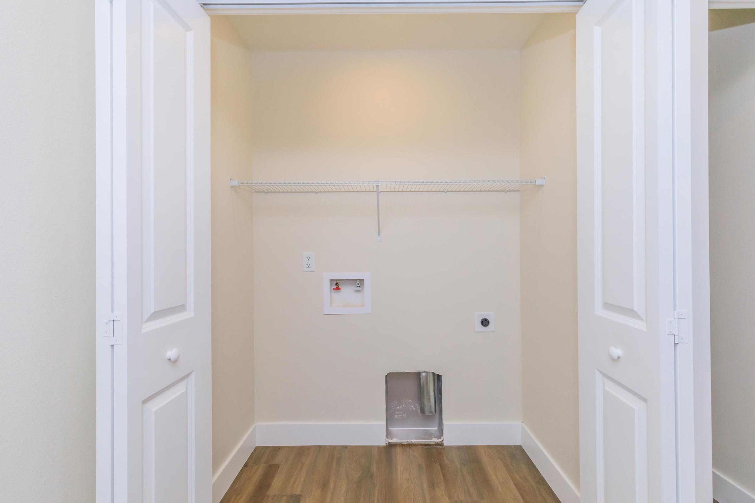 A spacious, empty laundry room featuring a shelf for storage, a wall-mounted electrical outlet, and connections for a washer and dryer. The room has light-colored walls and a warm wood-like floor, with double doors that open to reveal the space.