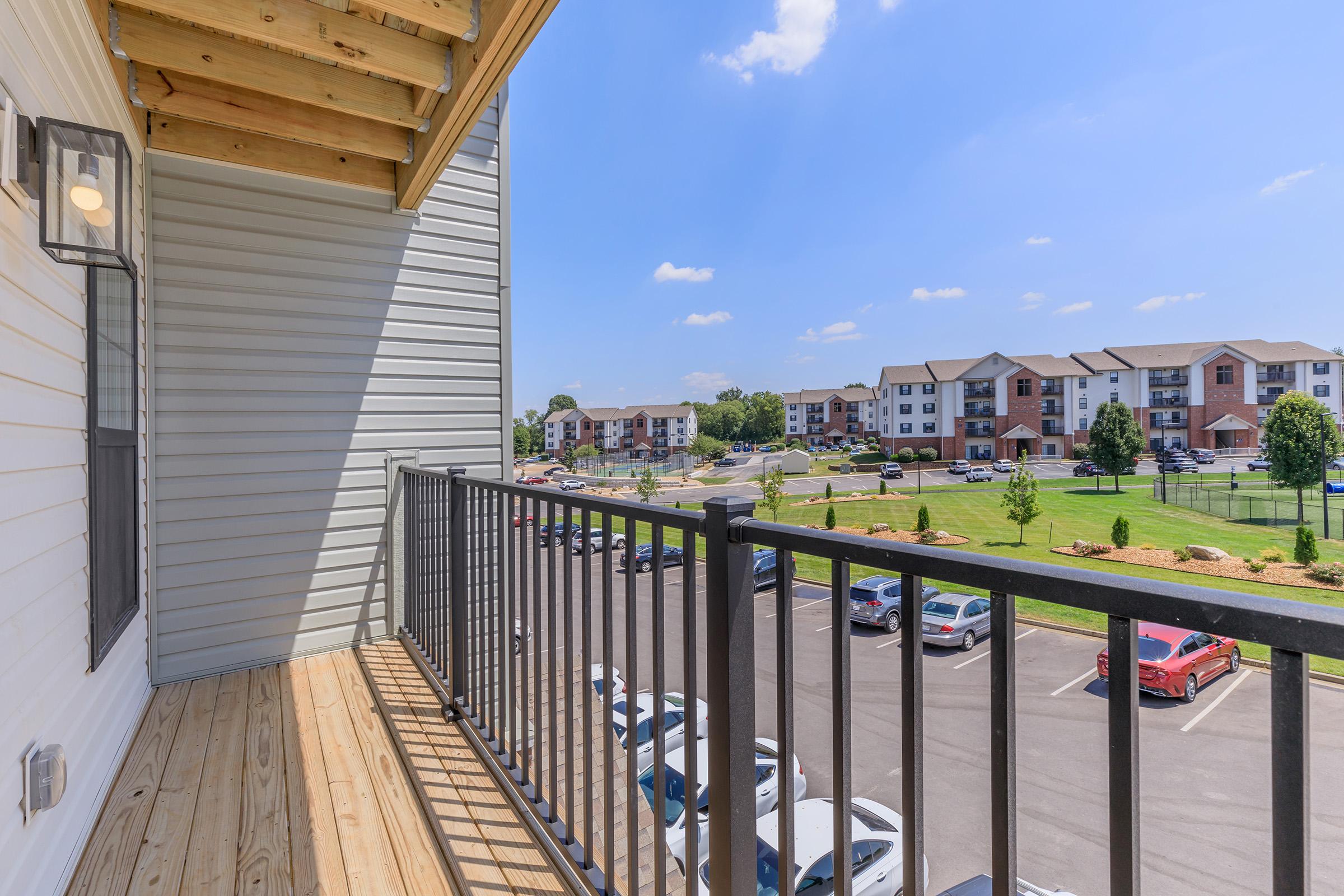 View from a balcony overlooking a parking lot and landscaped area with several apartment buildings in the background. The sky is clear and blue, with a few fluffy clouds, and the scene captures greenery and outdoor spaces, creating a tranquil residential atmosphere.