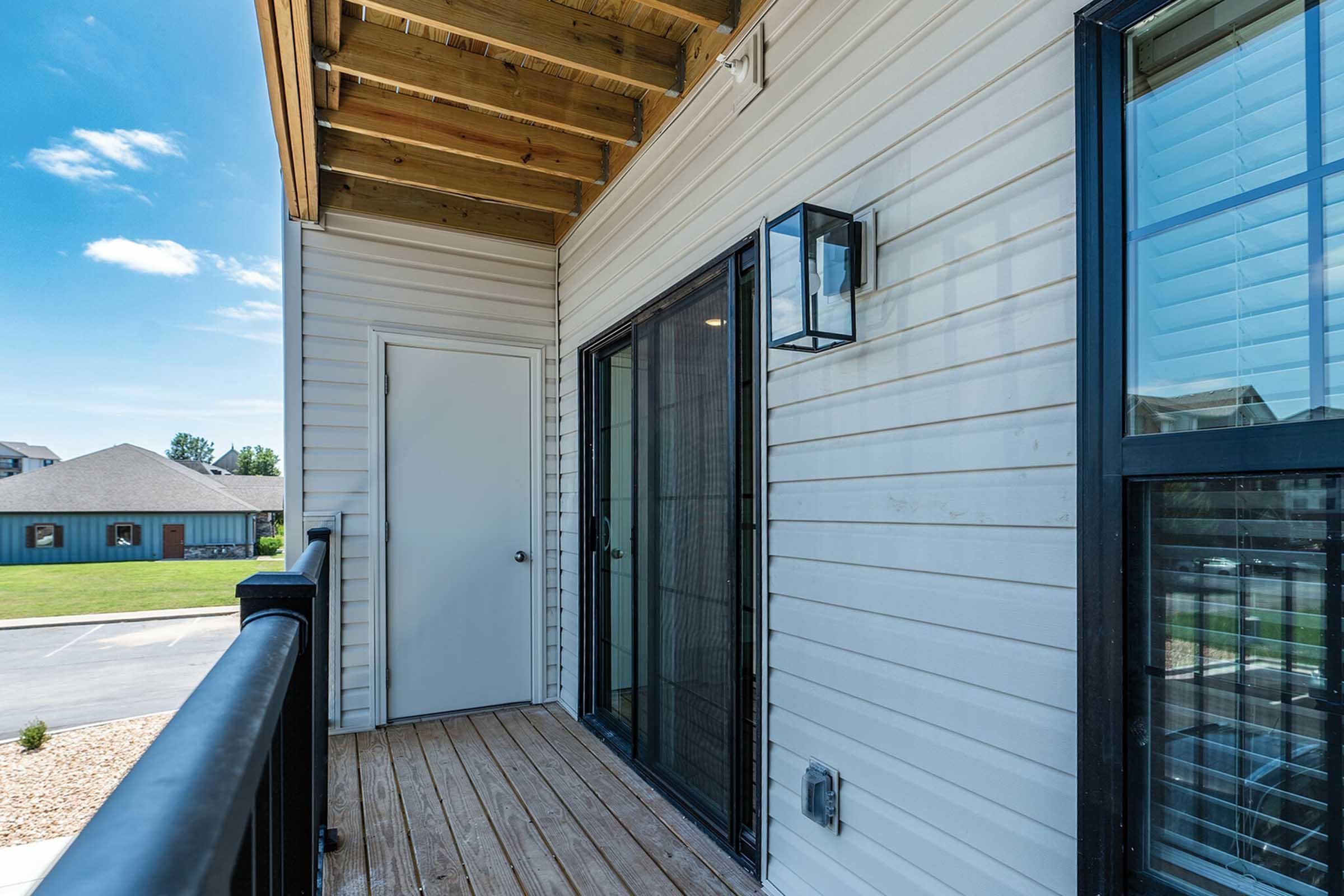 A view of a covered balcony with wooden flooring, featuring sliding glass doors, a white wall, and a lantern-style light fixture. The surrounding area includes a grassy space and nearby buildings in the background under a clear blue sky.