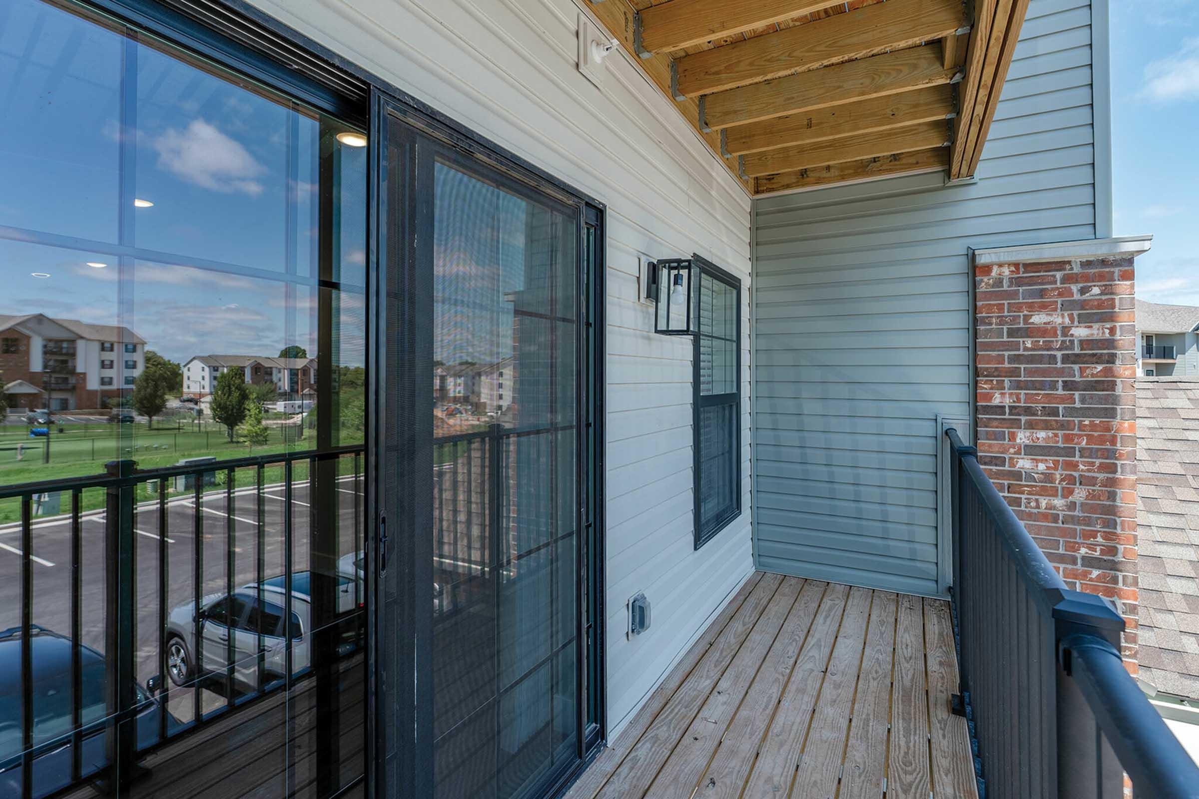 A view of a balcony with wooden flooring and a glass door leading inside. The railing is black, and the scene outside includes green grass and parking spaces. The sky is partly cloudy, indicating a bright day.
