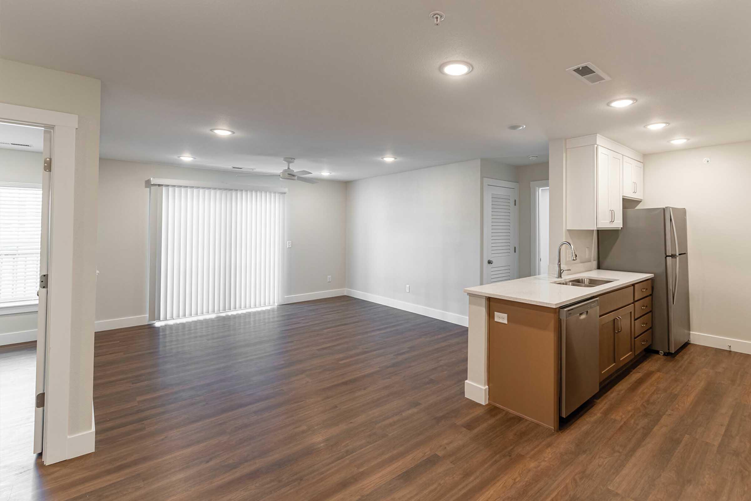 An interior view of a modern apartment living area with light-colored walls and hardwood flooring. The kitchen features light cabinets, a stainless steel refrigerator, and a countertop. A large window with white blinds allows natural light into the space, and there's a ceiling fan for ventilation.