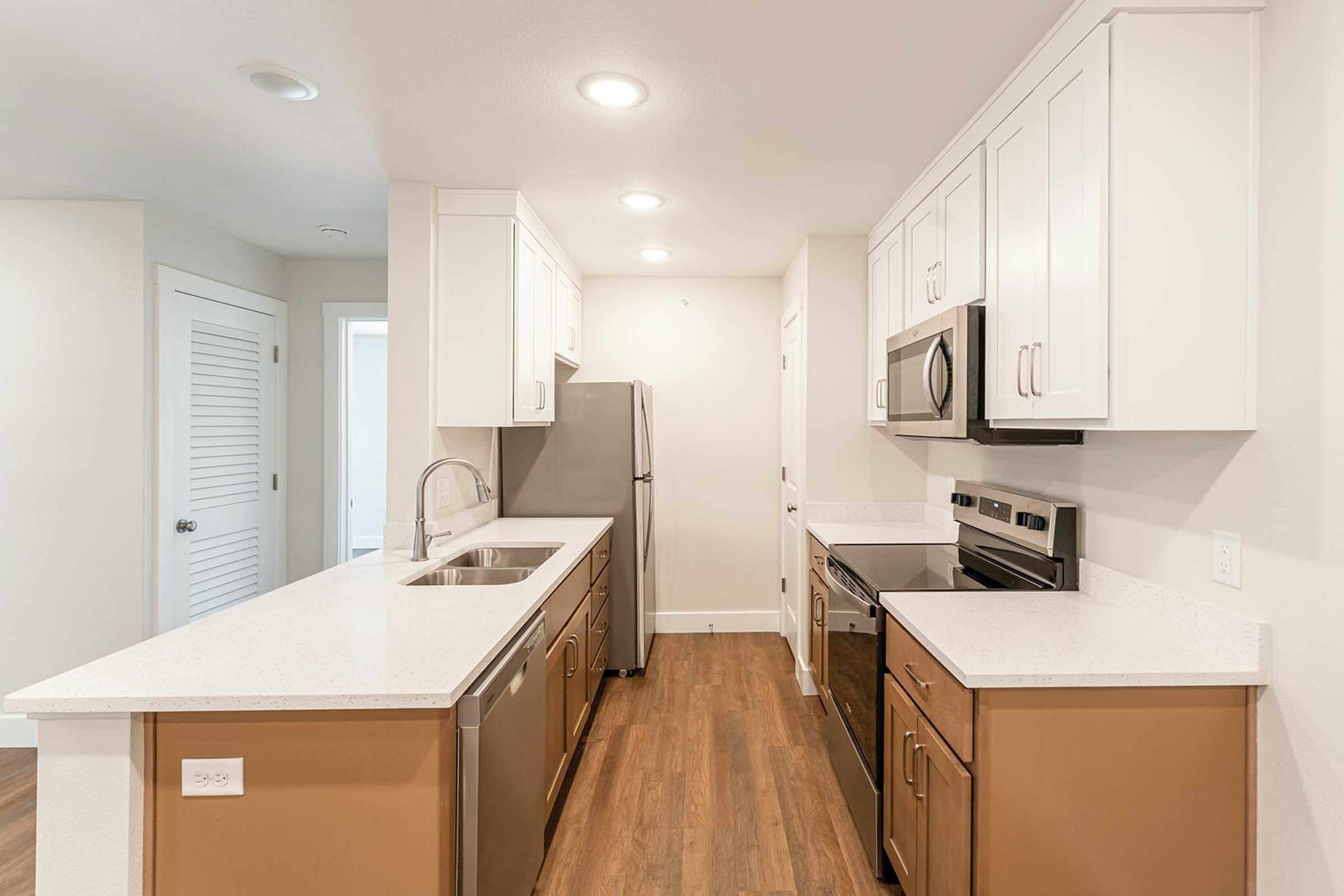 A modern kitchen featuring white cabinets, stainless steel appliances, and a combination of white and brown cabinetry. The countertops are light-colored, and the flooring is wood-like. Soft overhead lighting enhances the spacious feel of the room, with a view of an adjoining area.