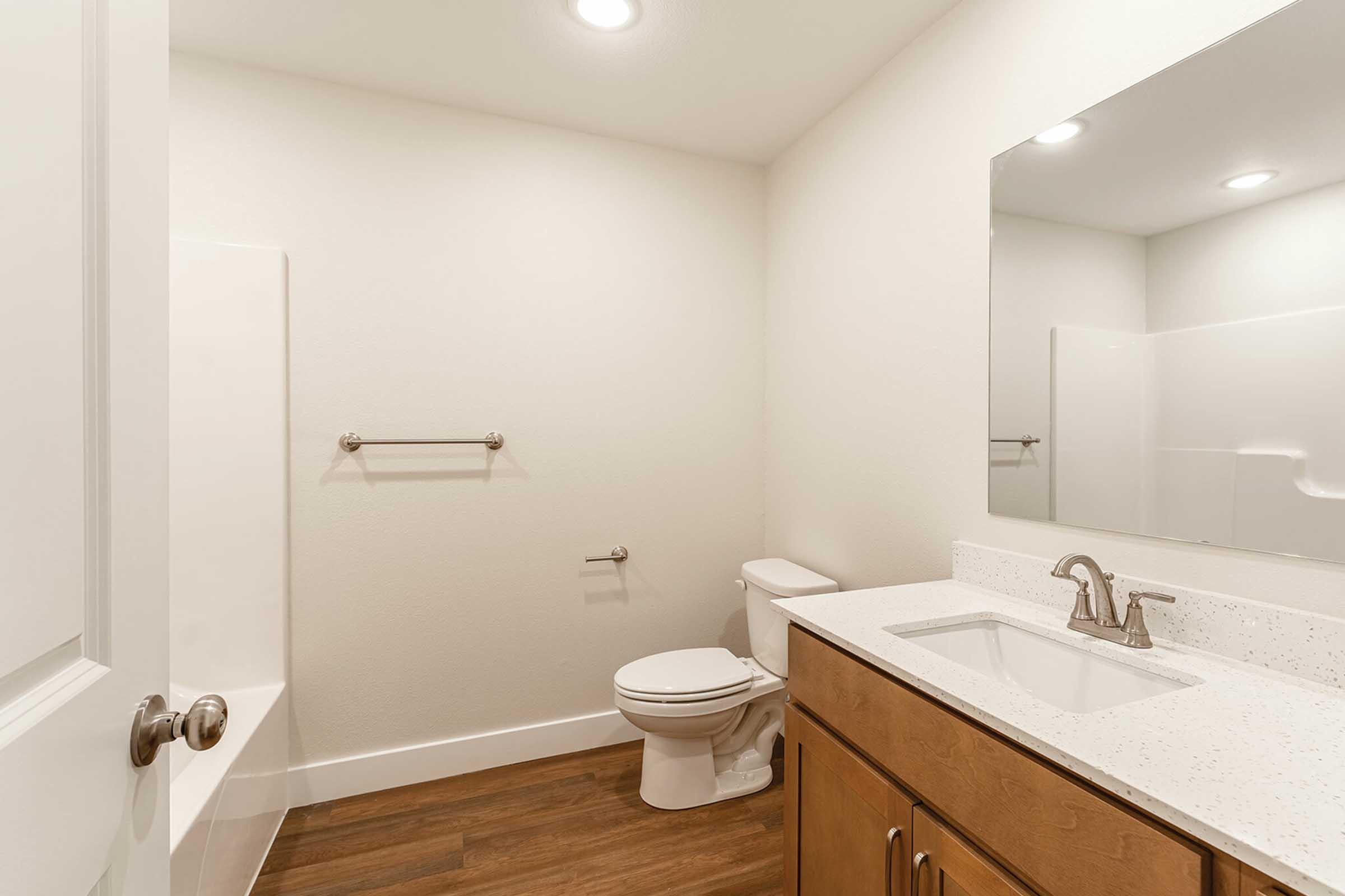 A modern and minimalistic bathroom featuring a white bathtub, toilet, and a vanity with a sink. The walls are painted a light color, and there is a large mirror above the sink. The flooring is wood-like, creating a warm atmosphere. Soft lighting enhances the overall bright and clean look of the space.