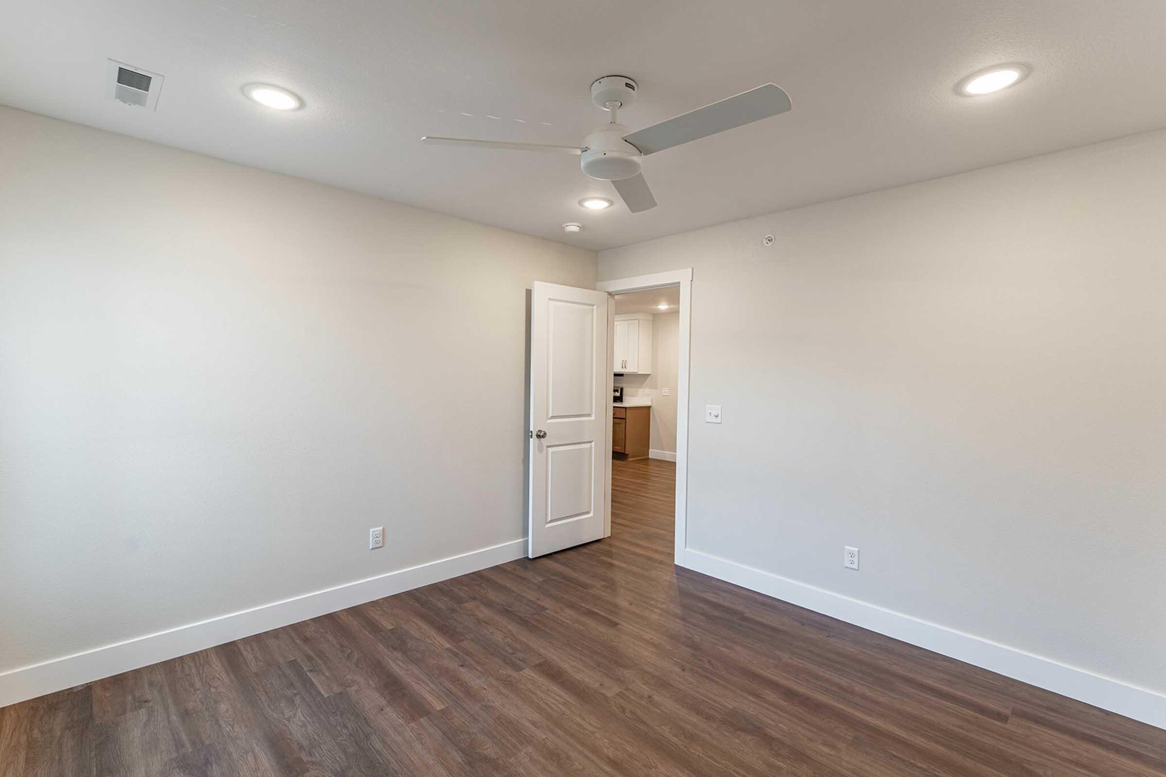 Interior view of an empty room featuring light wood flooring, white walls, and recessed lighting. A ceiling fan is mounted on the ceiling. A closed door is visible, leading to another area, possibly a kitchen, seen in the background. The space is bright and minimalistic, ideal for various uses.