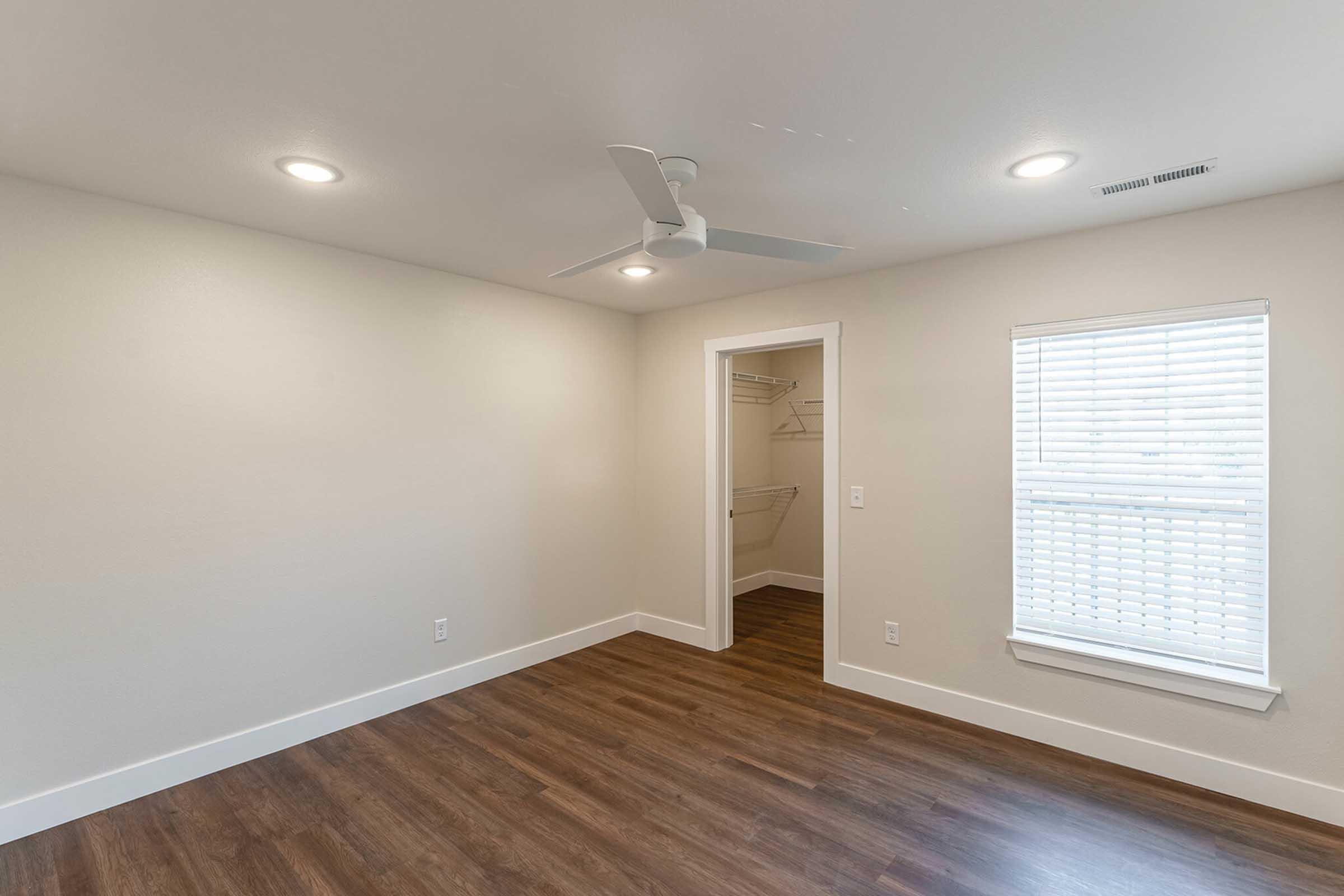 A bright, empty room featuring a ceiling fan, neutral-colored walls, and hardwood-style flooring. A closet is visible on one side with shelving, and a window with blinds lets in natural light. The overall atmosphere is clean and modern, suitable for various uses.