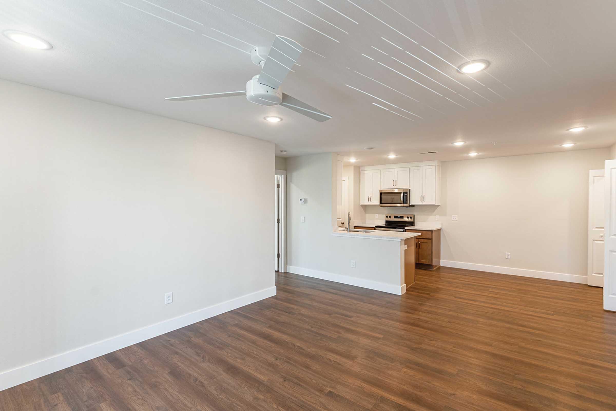 A modern, spacious living area featuring light-colored walls, a ceiling fan, and warm wood flooring. The kitchen is visible in the background, showcasing stainless steel appliances and white cabinetry. Ample natural light enhances the bright and inviting atmosphere.