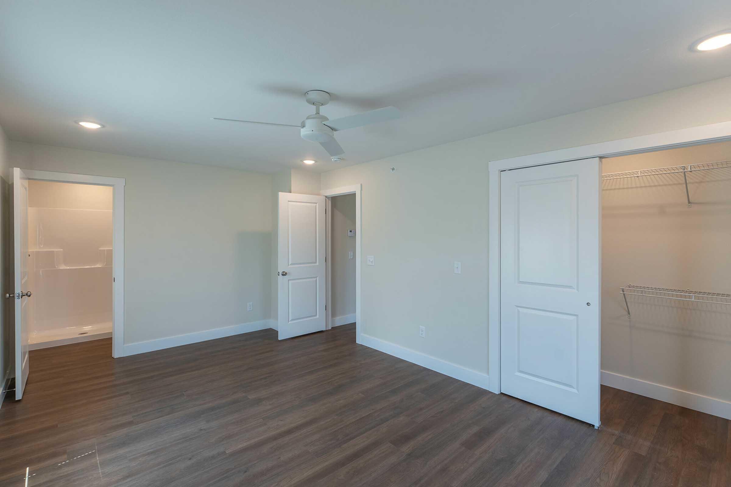 A spacious, empty bedroom with light-colored walls and a ceiling fan. The room features two doors: one leading to an adjoining space and the other to a closet with shelving. The floor is covered with modern laminate, and natural light is provided by recessed lighting and possible window space.