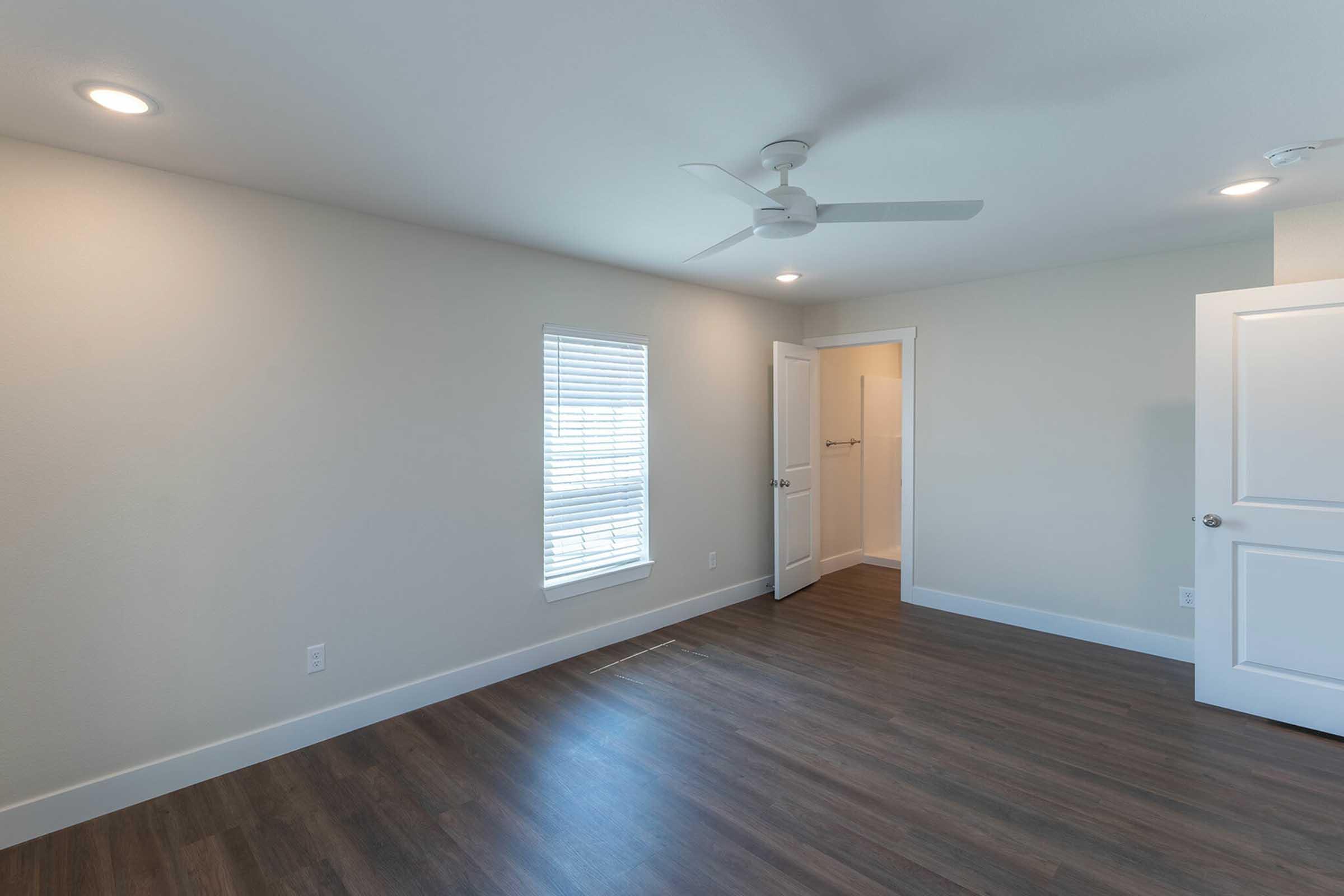 A bright, empty room with light-colored walls and brown wooden flooring. A window with blinds allows natural light to enter, and there's a ceiling fan installed. Two doors are visible, one leading into a closet or bathroom area, while the other appears to exit the room.