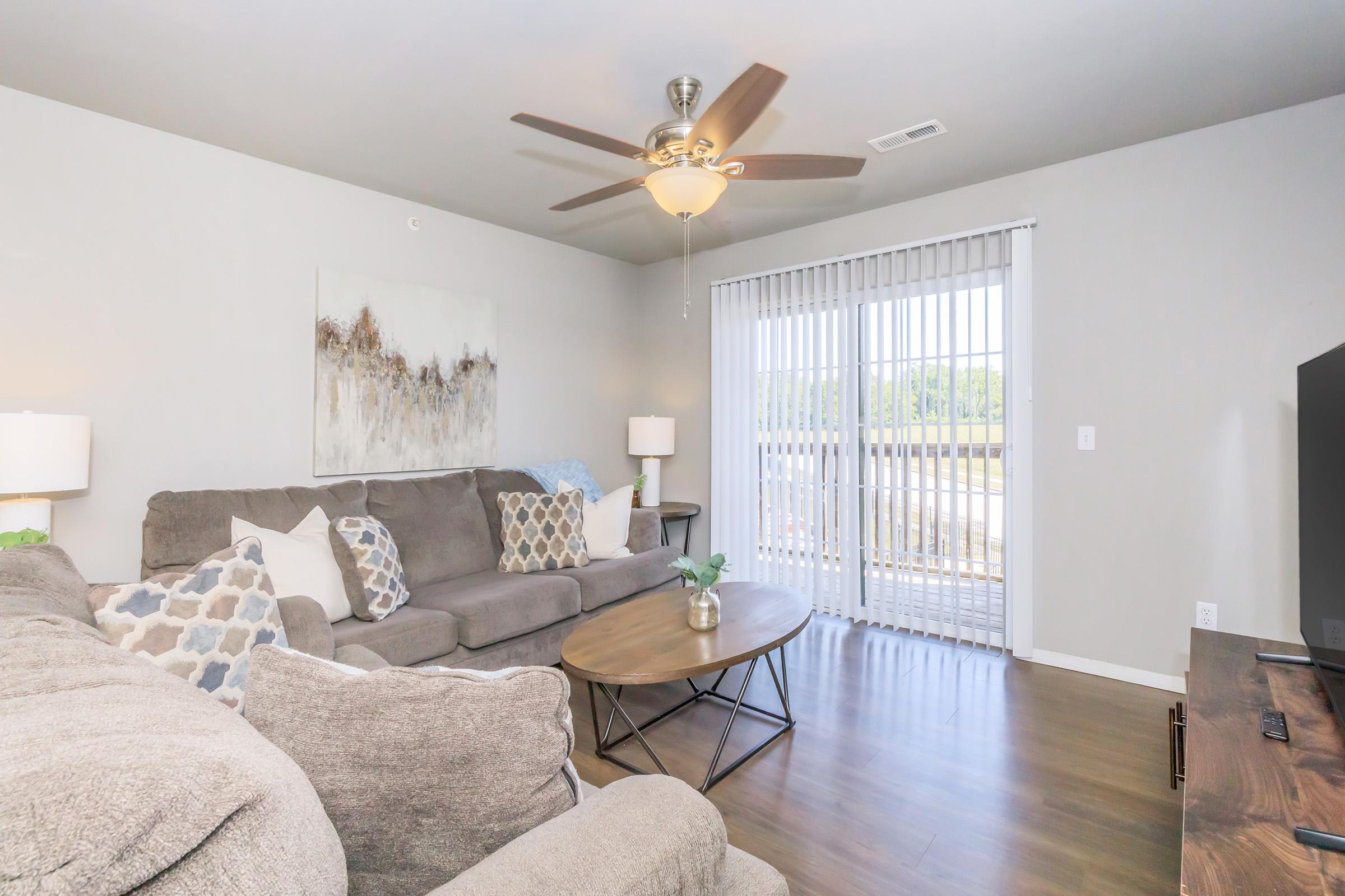 A cozy living room featuring a large gray sectional sofa with decorative pillows, a round coffee table, and a TV stand. The room has a ceiling fan and large windows with blinds, allowing natural light to brighten the space. A decorative wall art piece adds a touch of style to the neutral-colored walls.