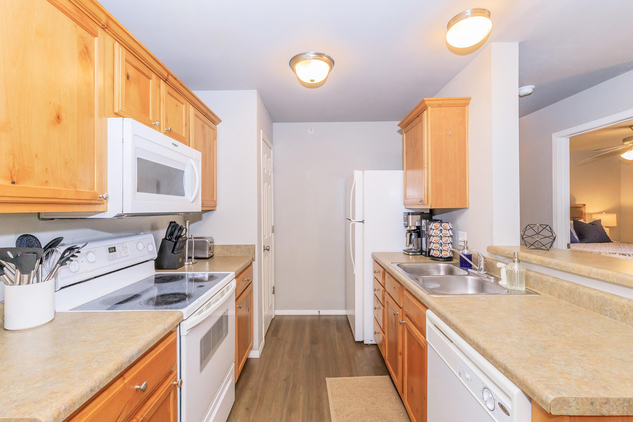 A modern kitchen featuring wooden cabinets, a refrigerator, stove, microwave, and sink. The countertops are light-colored, with a small rug on the floor. There are kitchen utensils and appliances visible, creating a clean and functional cooking space.