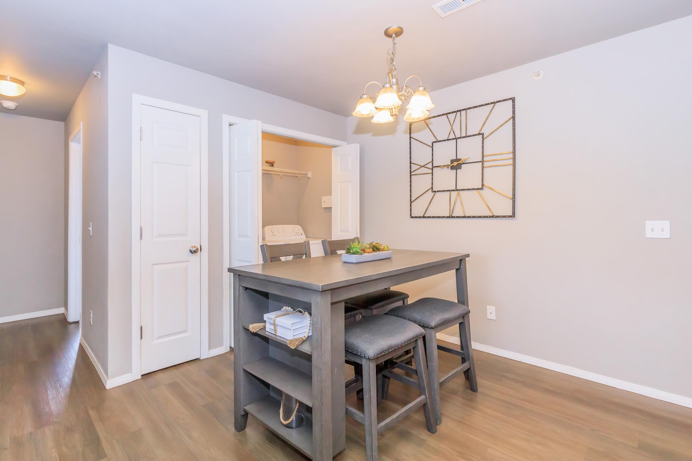 A modern dining area featuring a wooden table with high-backed stools, a decorative wall clock, and a light fixture above. The walls are painted in a soft gray, and there is a door leading to a laundry area in the background. The space is well-lit and tidy, creating a welcoming atmosphere.