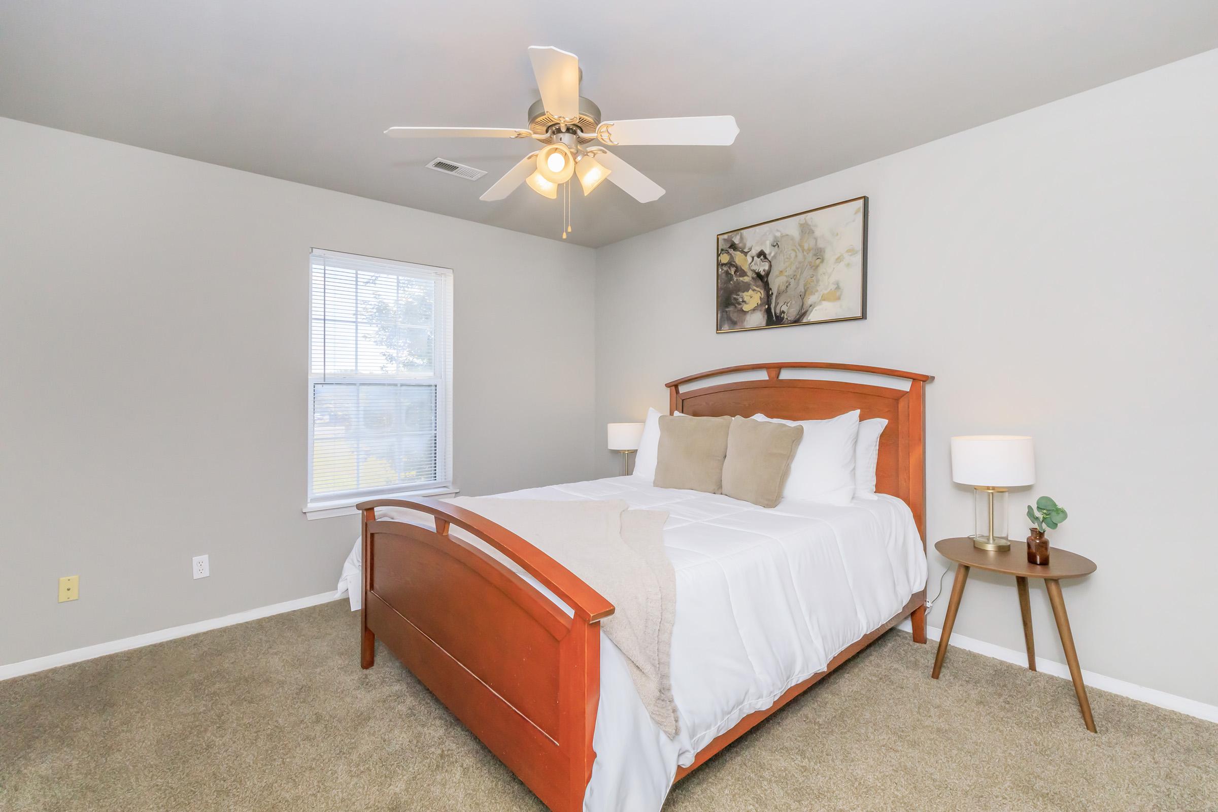 A well-decorated bedroom featuring a wooden bed frame with white bedding and decorative pillows. A ceiling fan is installed above, and there are two bedside tables with lamps. A piece of art hangs on the wall above the bed. Natural light comes through a window, illuminating the cozy, neutral-toned space.