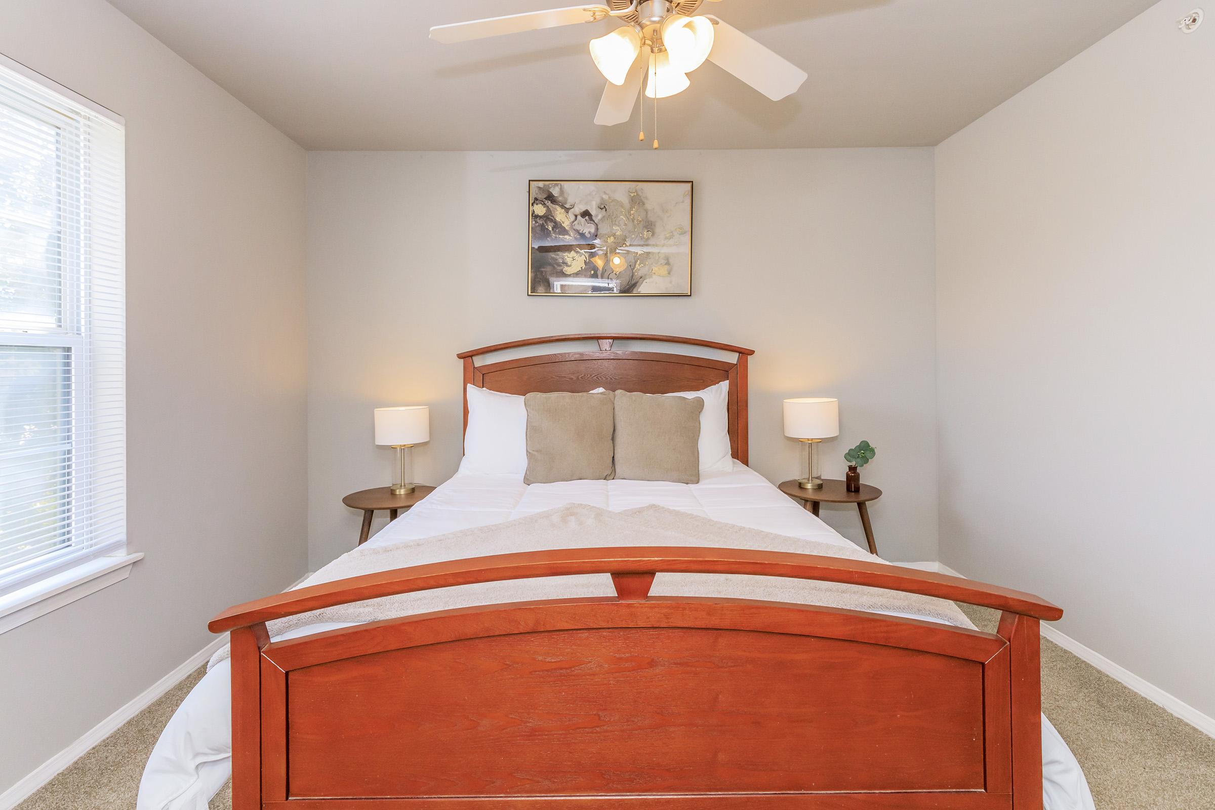 A cozy bedroom featuring a wooden bed with white bedding and two decorative pillows. Nightstands with lamps are on either side of the bed. A piece of artwork hangs above the bed, and a small plant is on one nightstand. Natural light comes through a window, creating a warm atmosphere.