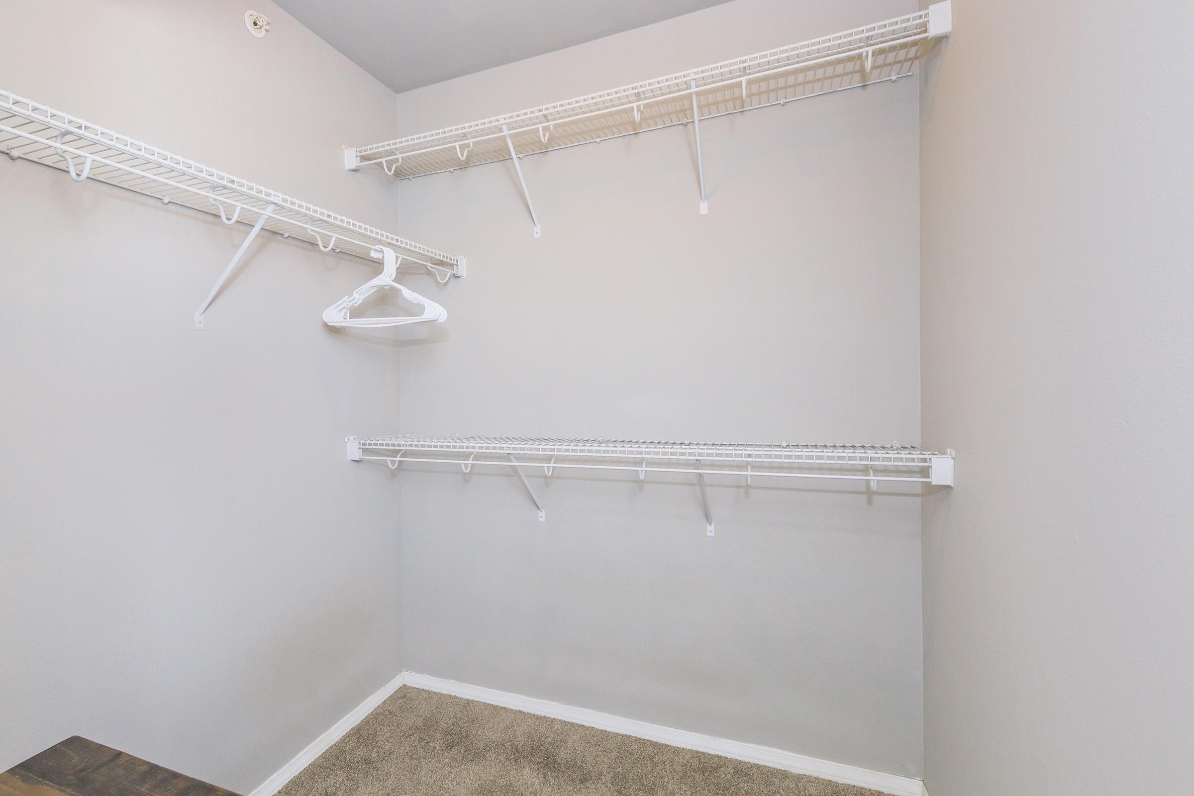 A spacious closet with beige carpeting and light gray walls. There are white wire shelves along the walls, with a single white hanger hanging from one of the shelves. The closet is empty, providing ample storage space.
