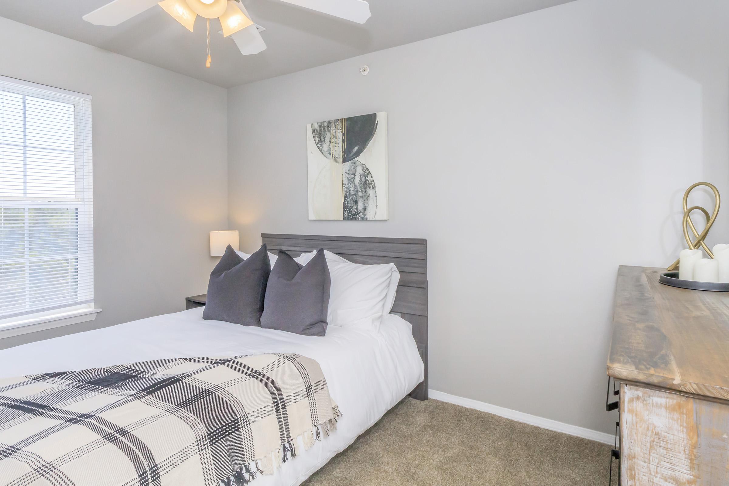 A cozy bedroom featuring a queen-sized bed with white linens and a plaid throw blanket. Two gray pillows rest at the head of the bed. A decorative wall art piece is hung above the bed, and a lampshade illuminates the space. A window allows natural light to enter, and a wooden dresser is positioned nearby.