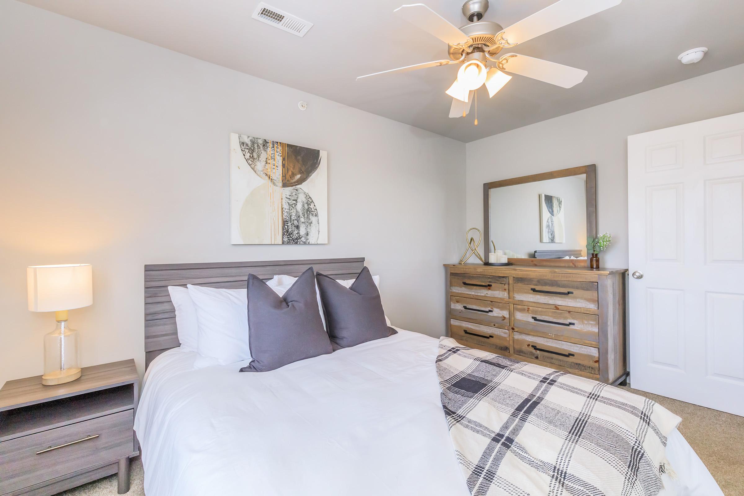 A cozy bedroom featuring a gray bed with decorative pillows, a wooden nightstand with a lamp, a dresser, a framed artwork on the wall, and a ceiling fan. The room has light gray walls and a warm, inviting atmosphere.