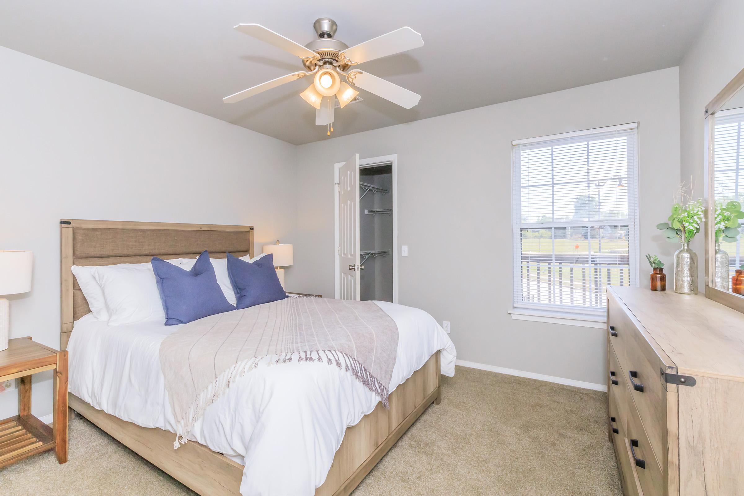 A cozy bedroom featuring a queen-sized bed with white linens and blue accent pillows. There are two bedside lamps on wooden nightstands, a dresser against the wall, and a ceiling fan. A window lets in natural light, revealing a view outside. The walls are painted in a soft gray tone, and the carpet is beige.