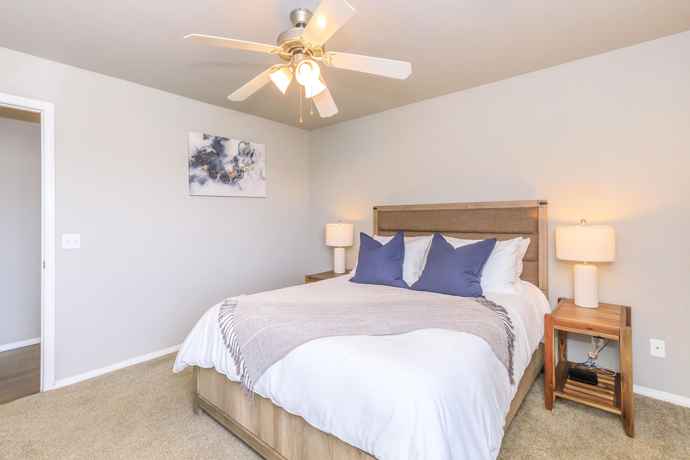 A cozy bedroom featuring a wooden bed with white bedding and blue accent pillows. Two bedside lamps provide soft lighting, and a ceiling fan hangs above. A piece of abstract art is displayed on the wall, and the room has neutral-colored walls with carpeted flooring. A door leads to another space.