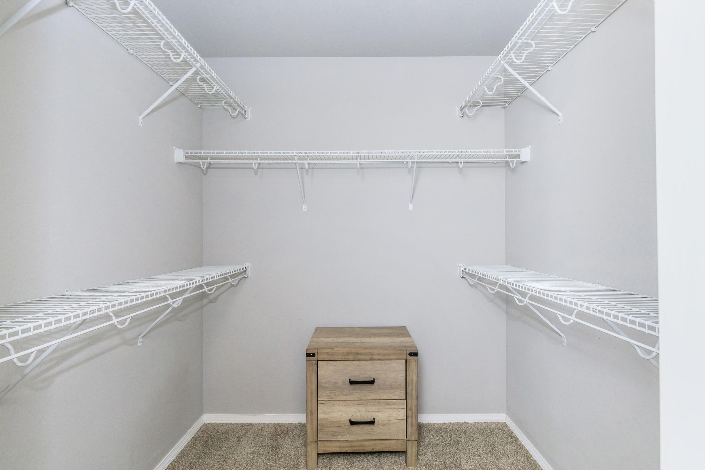 An empty closet with white wire shelves on both sides and a small wooden drawer unit in the center. The walls are painted a light color, and the floor is carpeted. The space appears clean and organized, suitable for storage or clothing.