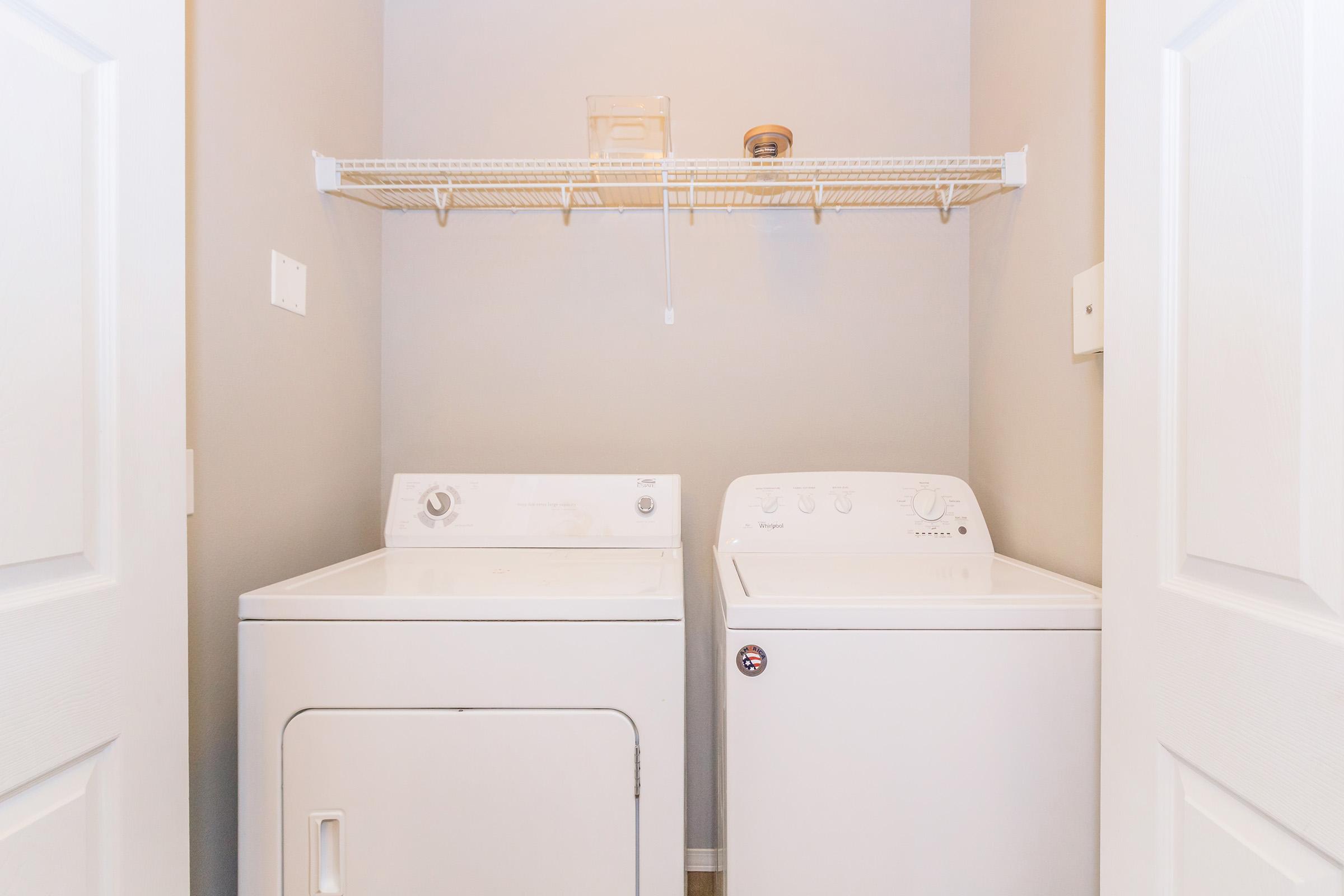 A clean laundry room featuring a white washer and dryer side by side. Above them, a wire shelf holds a small container. The walls are painted in a light gray color, and the room is well-lit, creating an organized and tidy appearance.