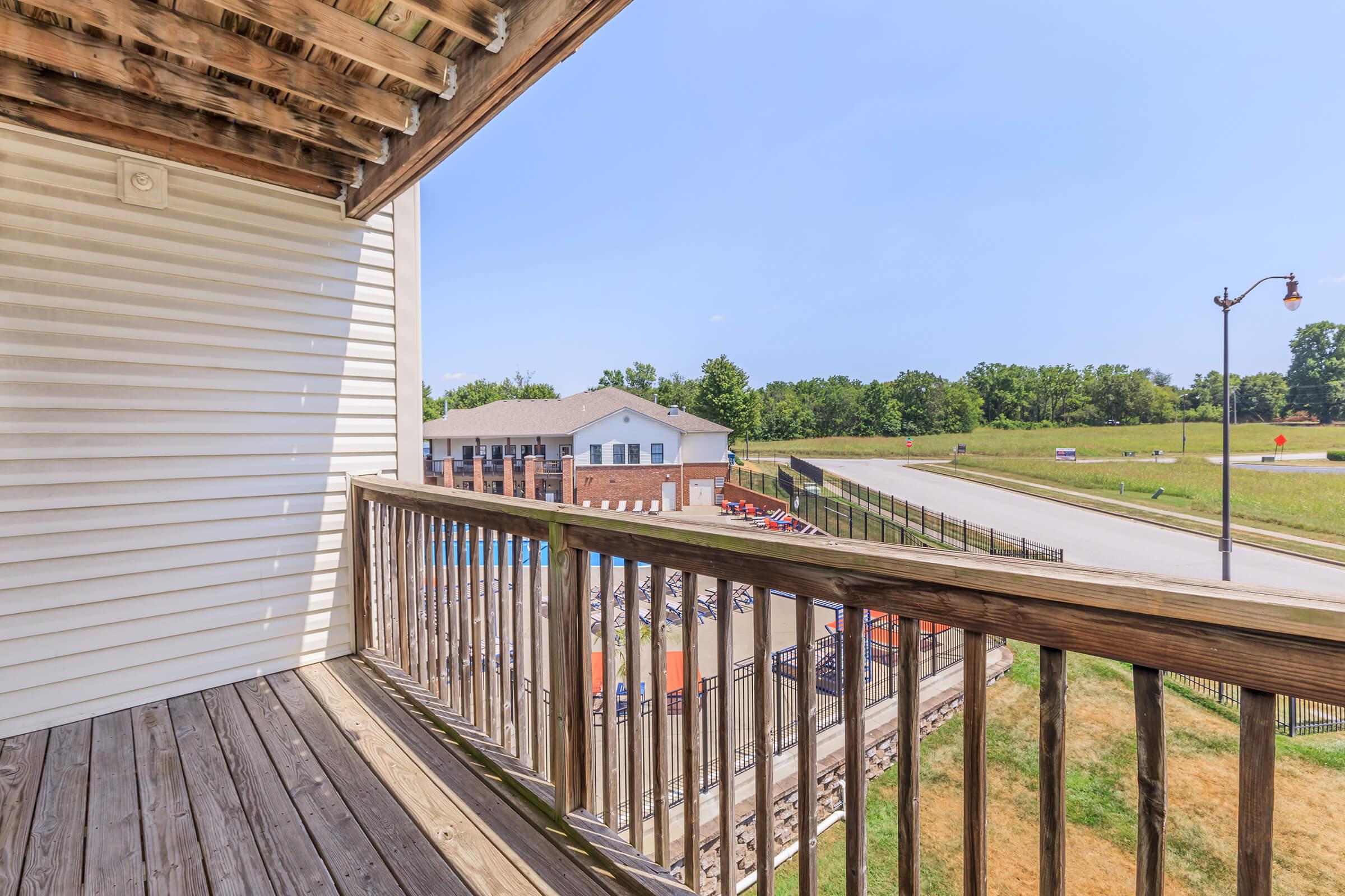 View from a balcony showing wooden railing and floor, overlooking a road and distant buildings. A swimming pool and outdoor area are visible below, surrounded by greenery and clear blue sky.