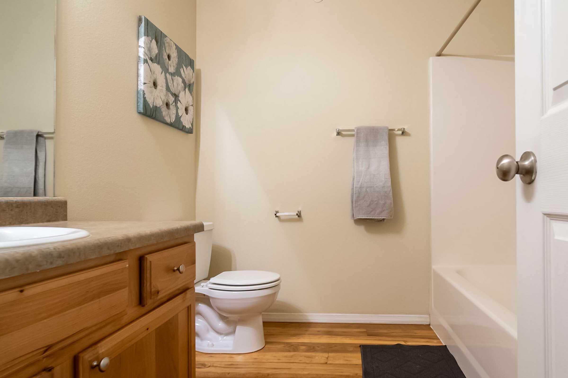 A clean and well-lit bathroom featuring a light-colored wall, a wooden vanity with a sink, a toilet, a gray towel hanging on a rack, and a white bathtub. Decorative floral artwork is on the wall, and the floor has a dark mat. The overall ambiance is neat and inviting.