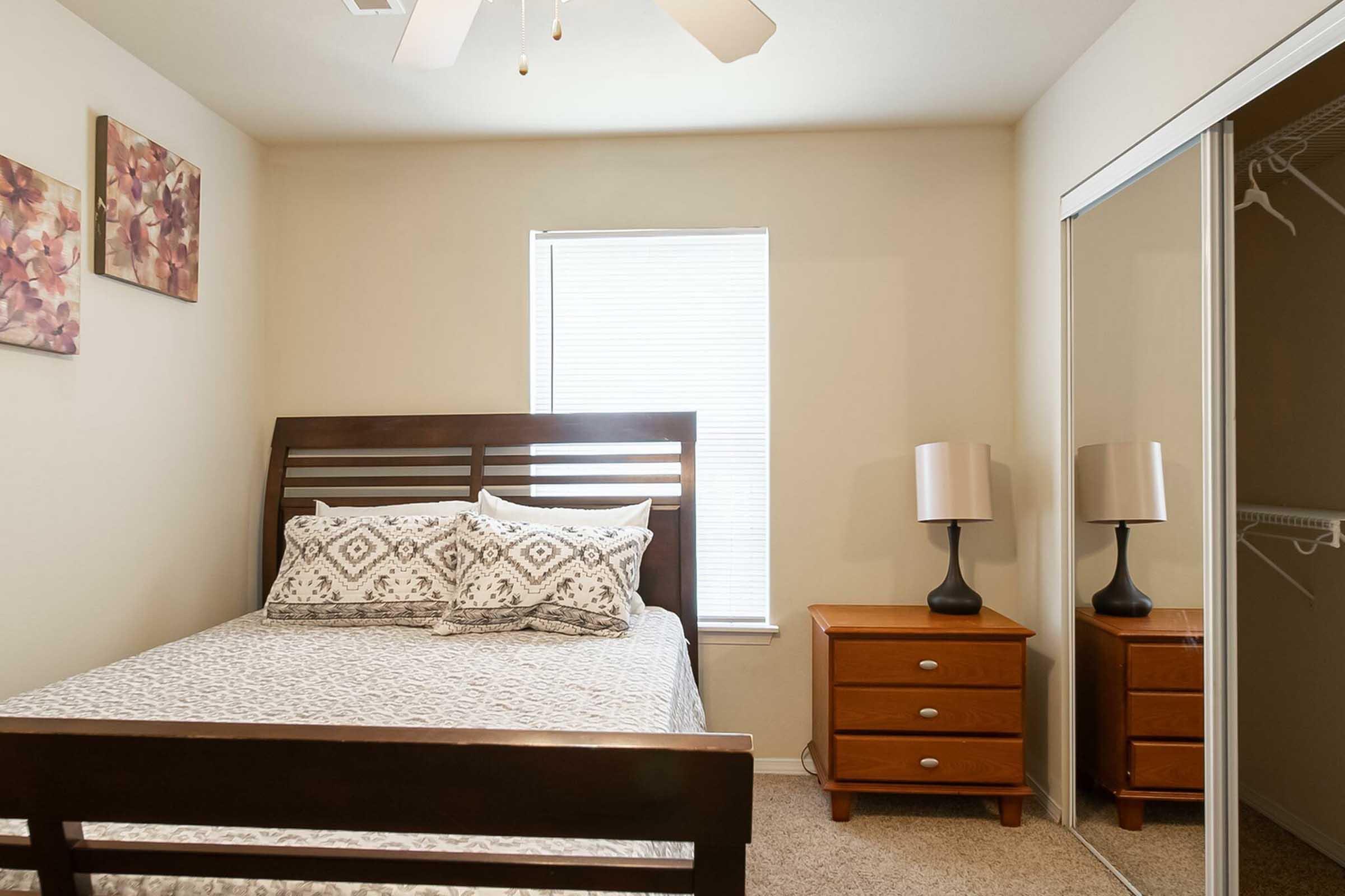 Cozy bedroom featuring a bed with a decorative gray and white patterned duvet and two matching pillows. A bedside table with a lamp stands next to the bed. The room has a large window with blinds, allowing natural light, and a mirrored closet door reflects the room's decor.