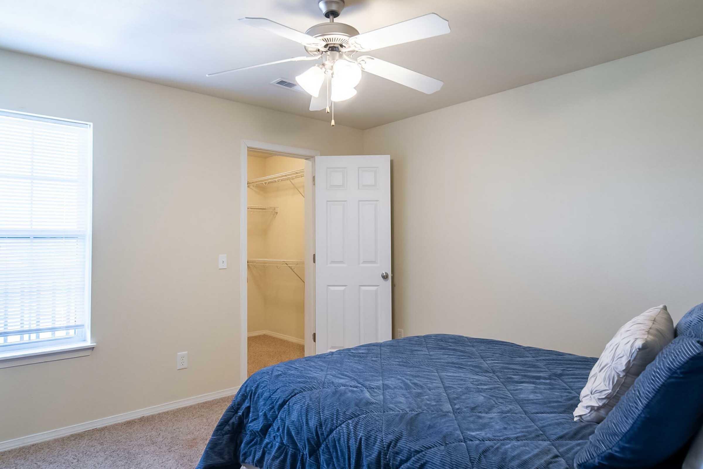 A well-lit bedroom featuring a blue bedspread, a ceiling fan, and beige walls. A closet door is partially open, revealing shelves inside. The room has a large window with blinds, providing natural light. The carpeted floor adds a cozy touch to the space.