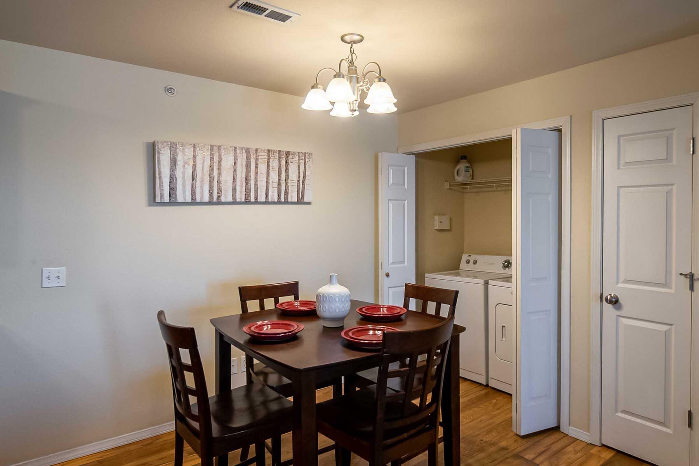 A cozy dining area featuring a dark wood table with four chairs, each set with a red plate. Nearby, there’s a door leading to a laundry room, and a light fixture with three bulbs. The wall is adorned with a decorative piece depicting trees. The flooring is wooden, creating a warm and inviting atmosphere.