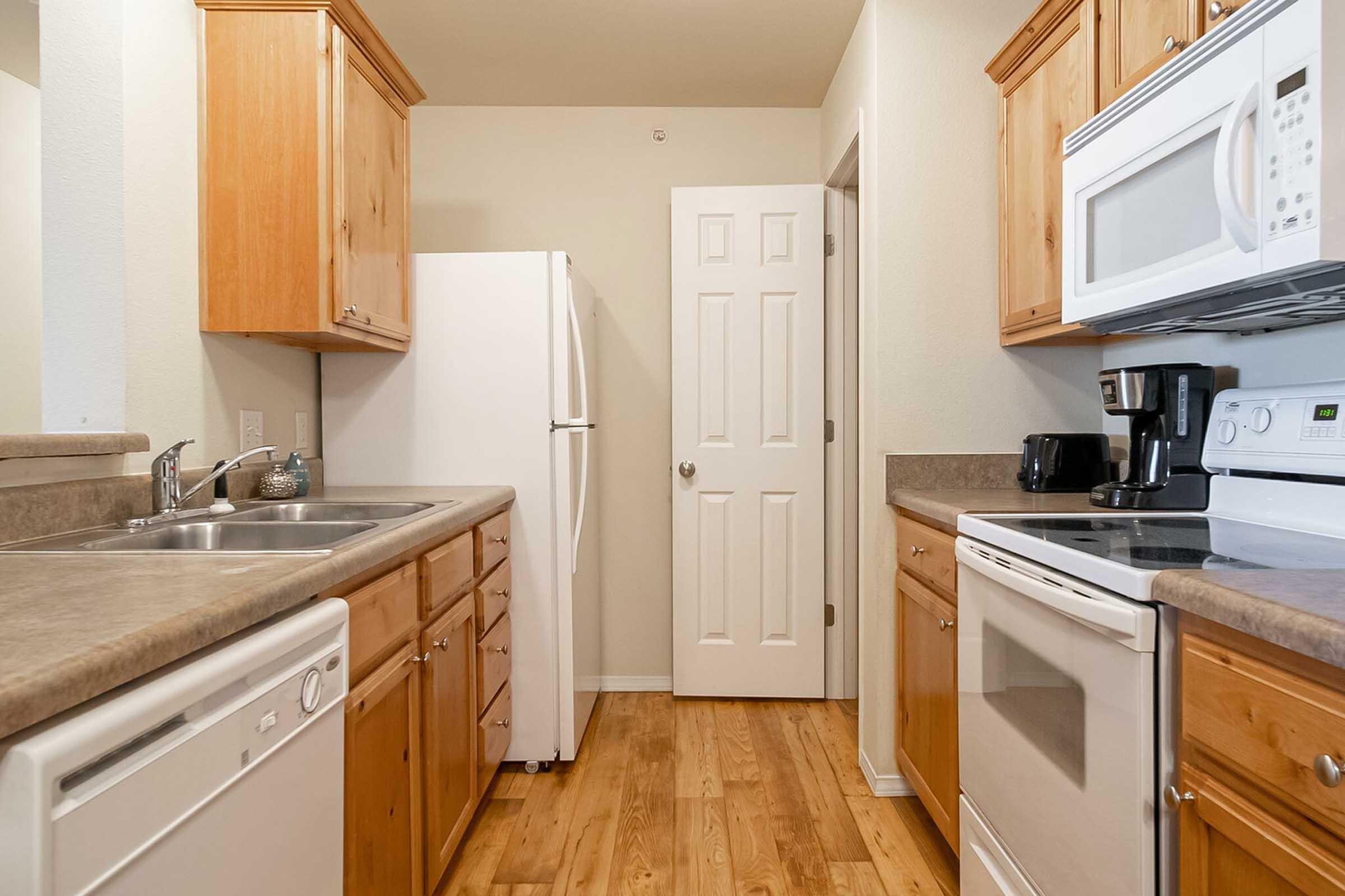 A modern kitchen featuring wooden cabinets, a double sink, a refrigerator, and various kitchen appliances including a microwave and coffee maker. The countertop is beige, and the flooring is light wood. A door is visible leading to another room. The overall design is clean and inviting.