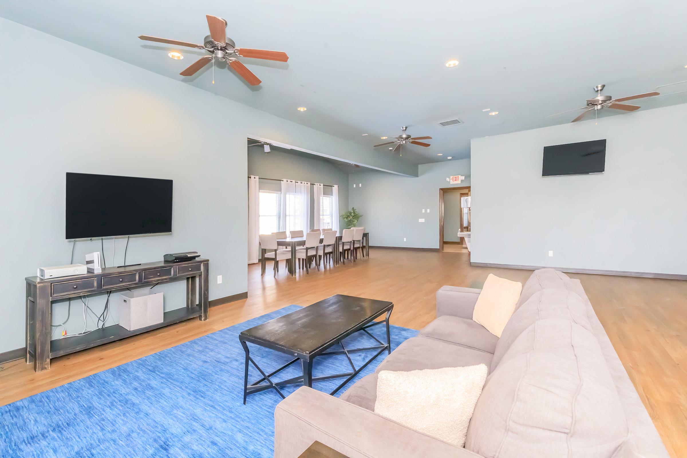 Spacious living area featuring a gray sectional sofa, a black coffee table, and a large blue area rug. There is a flat-screen TV mounted on the wall and ceiling fans above. In the background, a dining area is visible with a table and chairs, illuminated by natural light from windows.