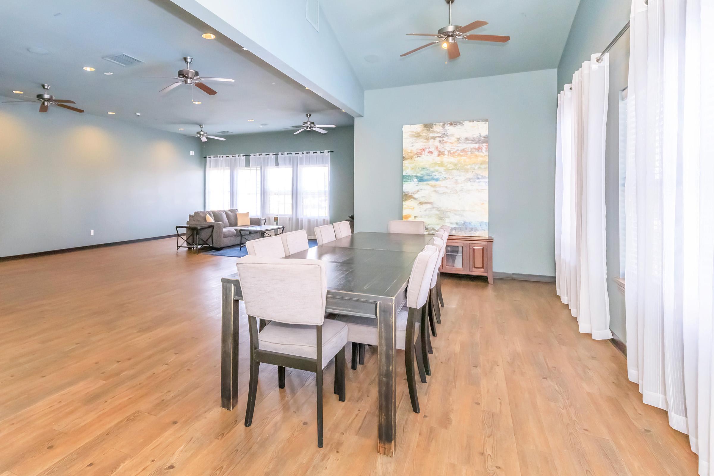 A spacious dining area featuring a long table with eight chairs, light wood flooring, and large windows adorned with white curtains. The room has ceiling fans and a large abstract painting on the wall, creating a modern and inviting atmosphere.