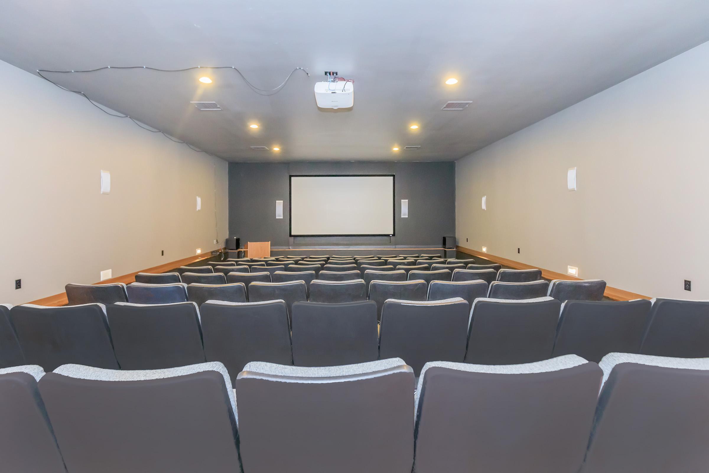 A spacious, empty cinema room with gray walls, arranged seating, and a large screen at the front. A projector is mounted on the ceiling, and the space is equipped with sound speakers along the walls. The atmosphere is quiet and ready for a film screening.