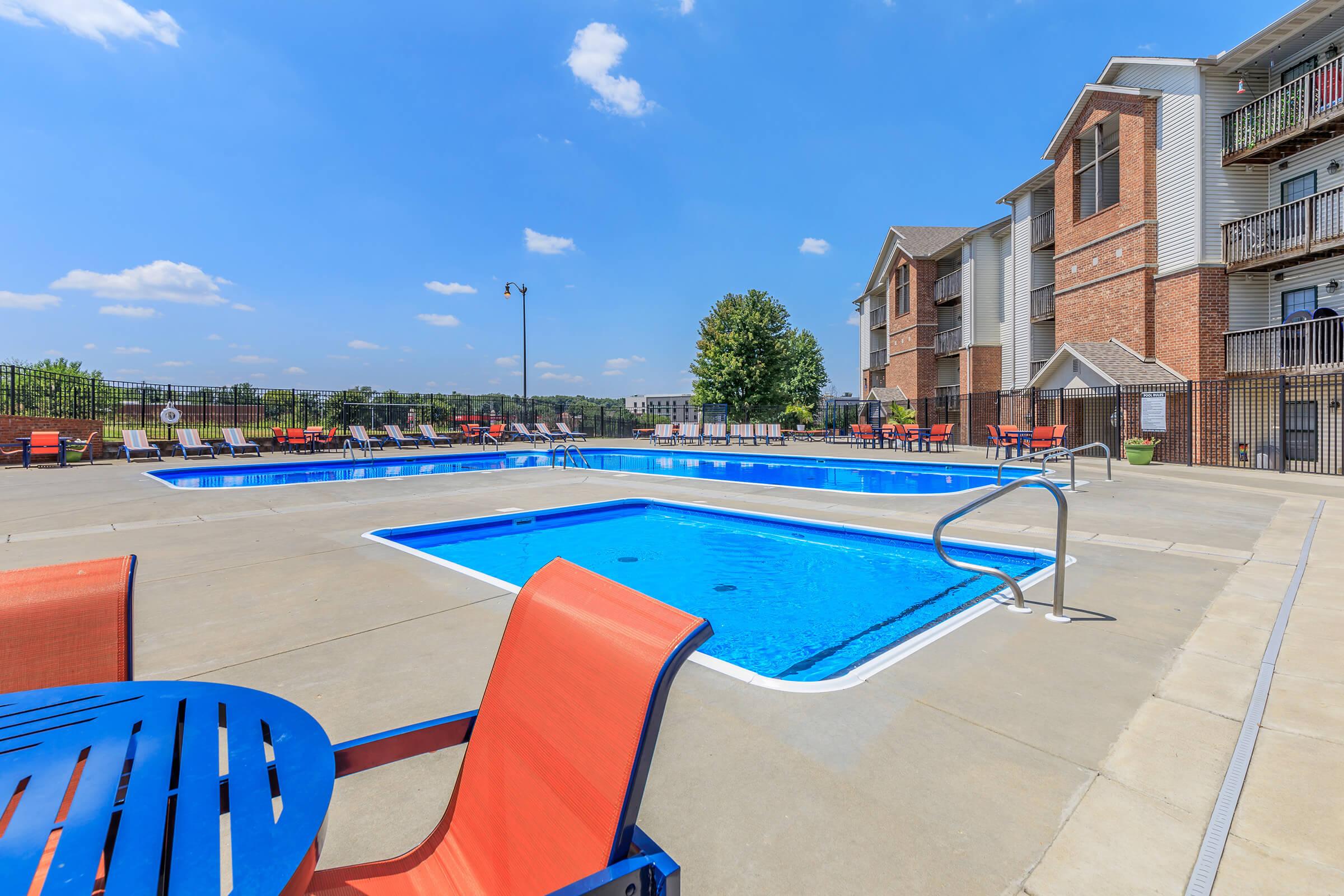 A bright outdoor pool area featuring two swimming pools, surrounded by lounge chairs and tables. The sky is clear with a few clouds, and there are residential buildings in the background. The pool deck is designed with a modern aesthetic, incorporating vibrant colors and comfortable seating.