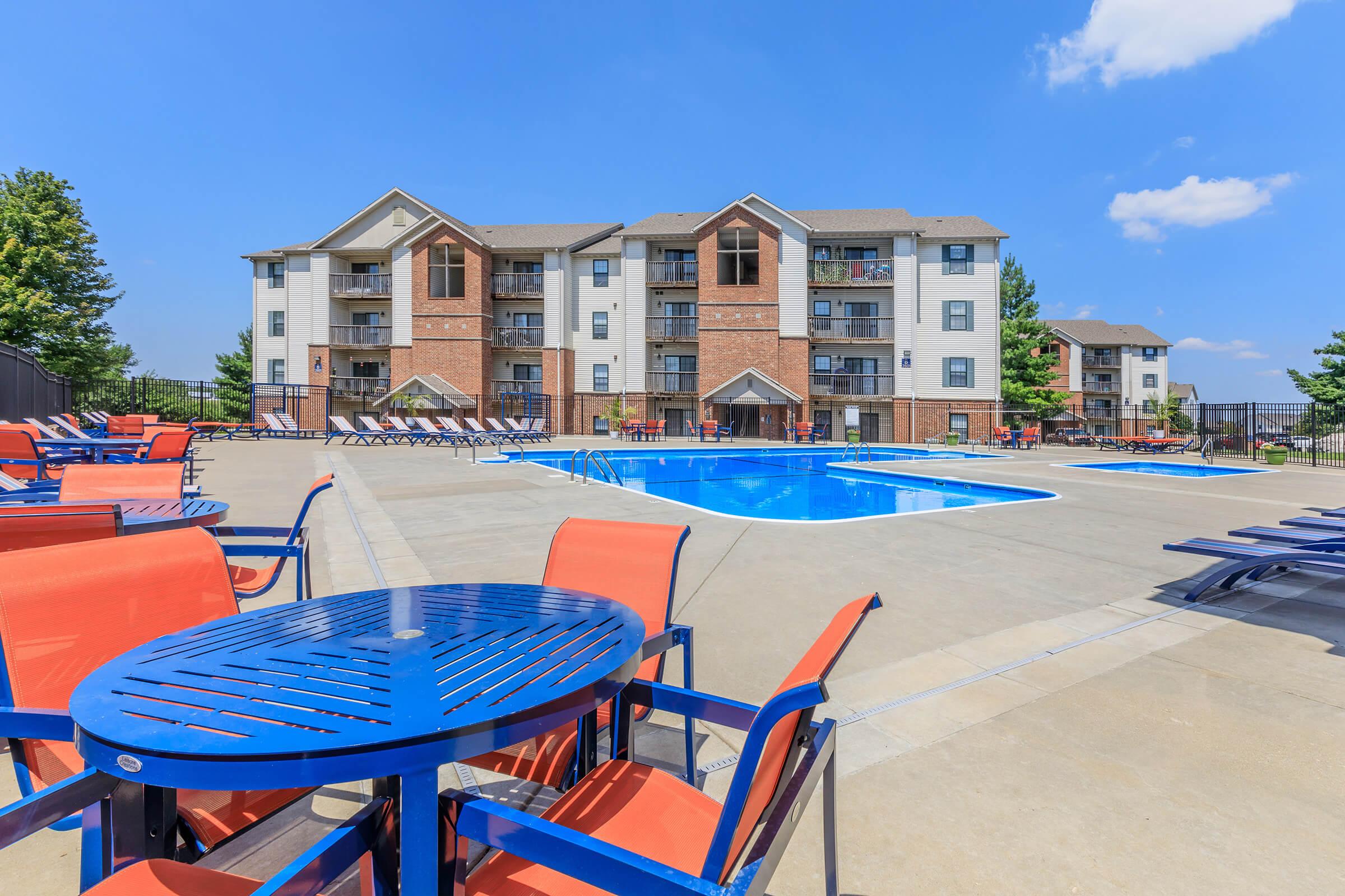 A vibrant outdoor pool area with a large swimming pool surrounded by lounge chairs and tables. In the background, a multi-story apartment building can be seen, set against a clear blue sky with a few trees nearby. The scene conveys a relaxing and inviting atmosphere.