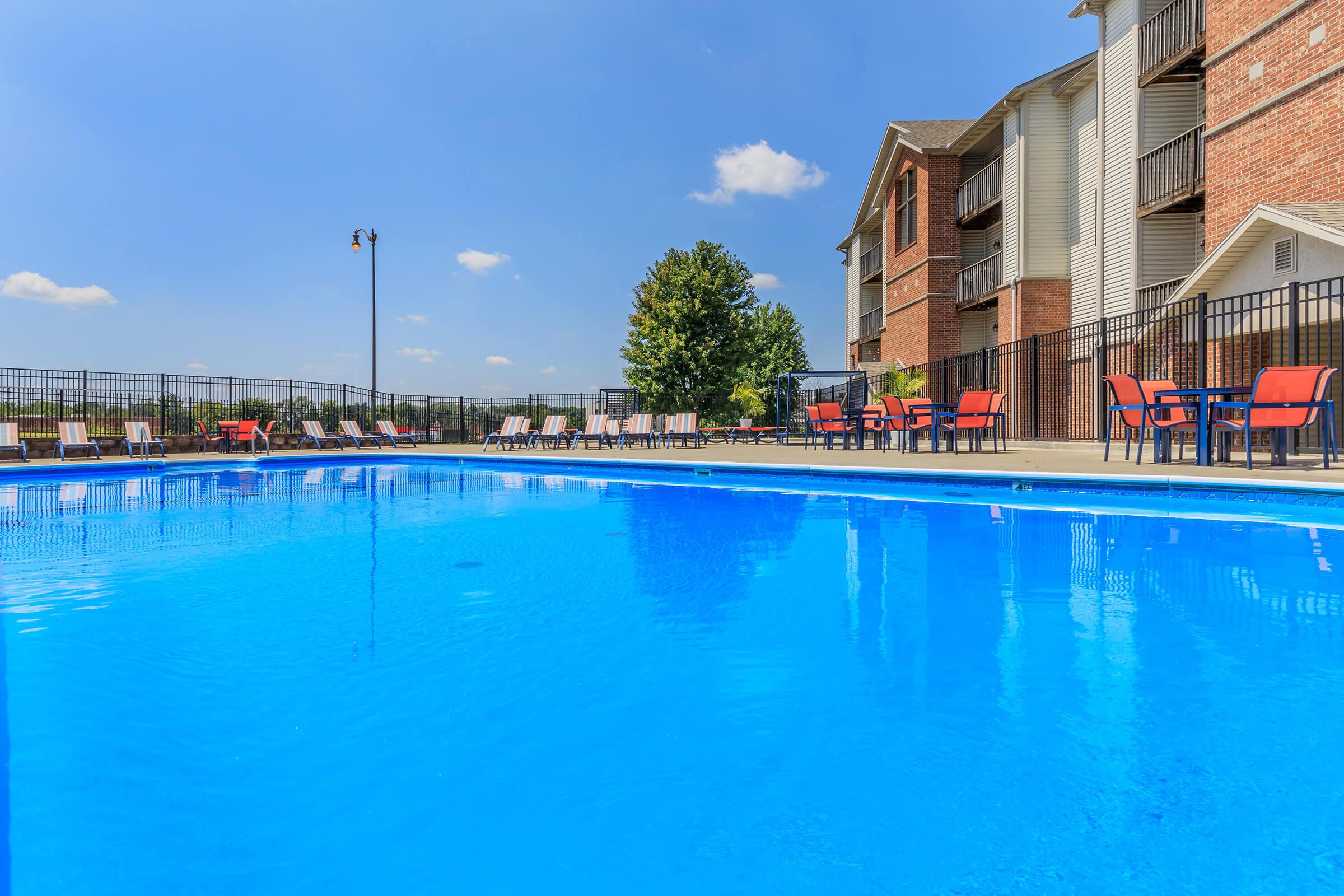 A bright blue swimming pool surrounded by lounge chairs and tables, with a clear sky overhead. Nearby, there are multi-story residential buildings visible in the background, creating a relaxing outdoor atmosphere for leisure activities.