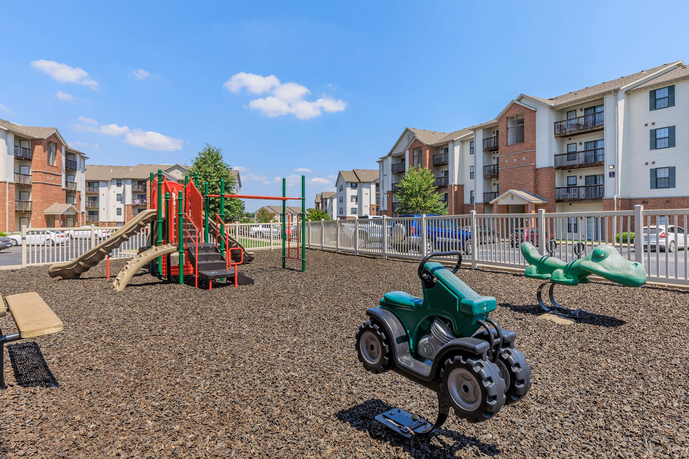 A playground area featuring a green tractor spring rider, a green alligator spring rider, and a colorful slide structure. The playground is surrounded by safety fencing and is situated near residential apartments under a clear blue sky.