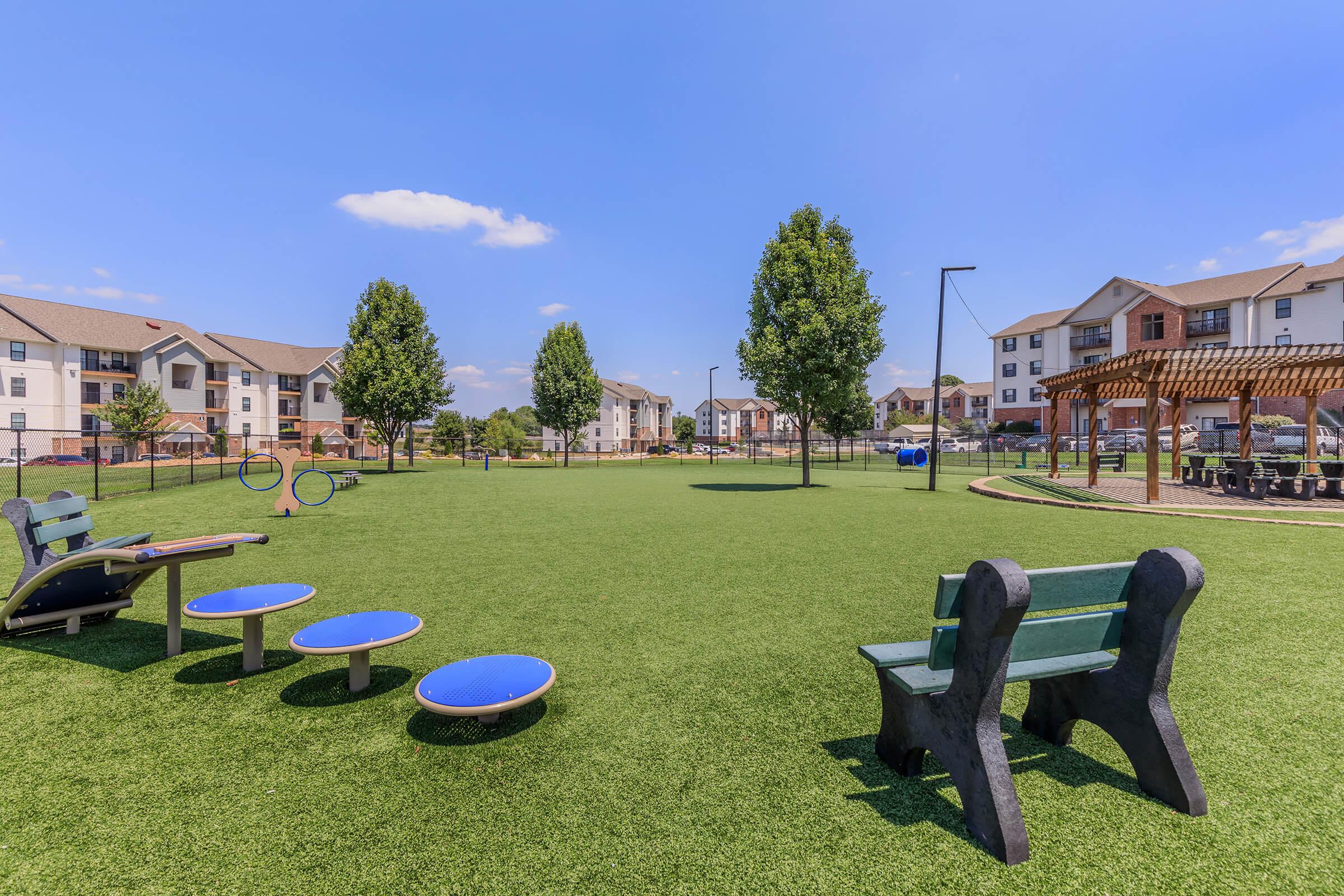 A spacious, well-maintained park featuring green artificial turf, benches, and circular seating. In the background, there are apartment buildings and a shaded pergola. The sky is clear with a few clouds, creating a bright and inviting atmosphere for outdoor activities.