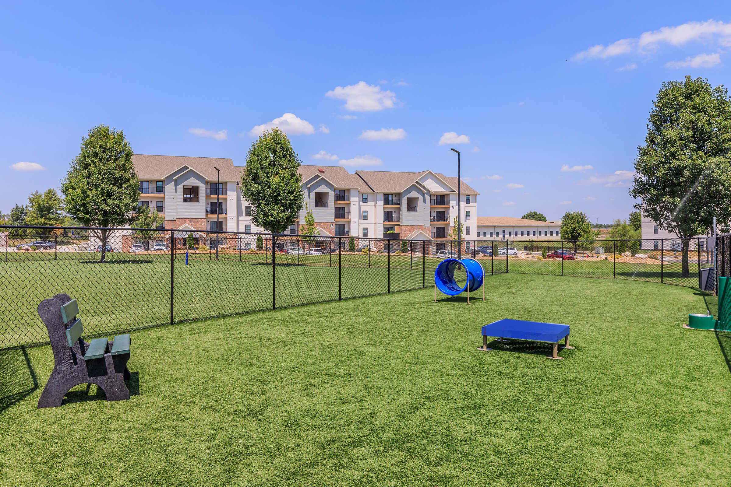 A landscaped dog park featuring a grassy area with benches, a tunnel, and a blue agility table. In the background, there are residential buildings under a clear blue sky with a few clouds. The park is enclosed by a chain-link fence, providing a safe space for dogs to play.
