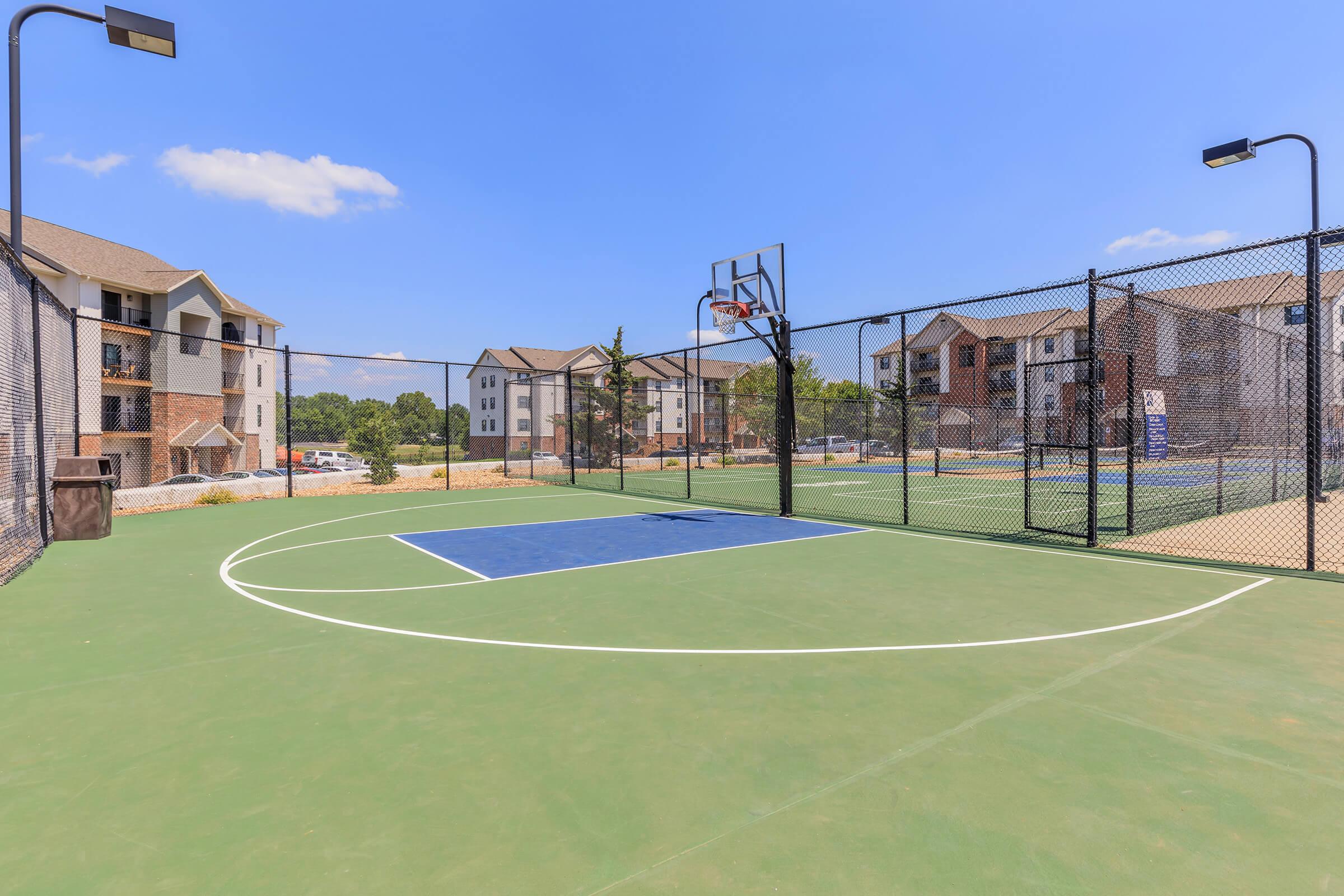 A well-maintained outdoor basketball court surrounded by a chain-link fence, with a blue three-point line, located near residential apartment buildings under a clear blue sky. The court is illuminated with black pole lights, providing a recreational area for residents.
